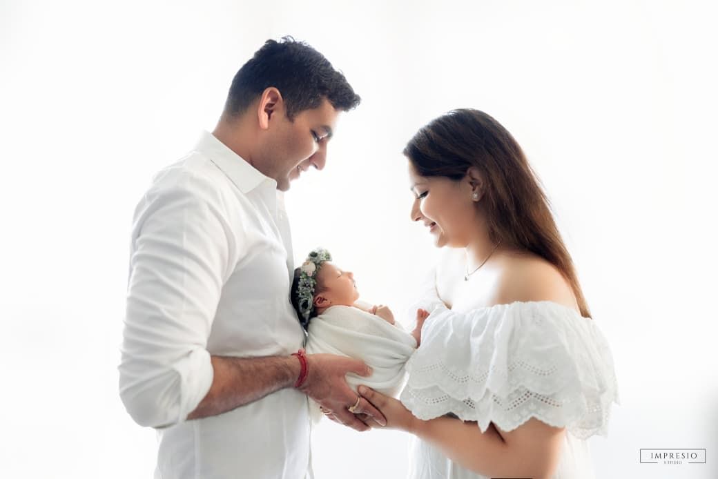 A couple in white outfits tenderly holds their newborn baby during a bright, airy newborn photography session.