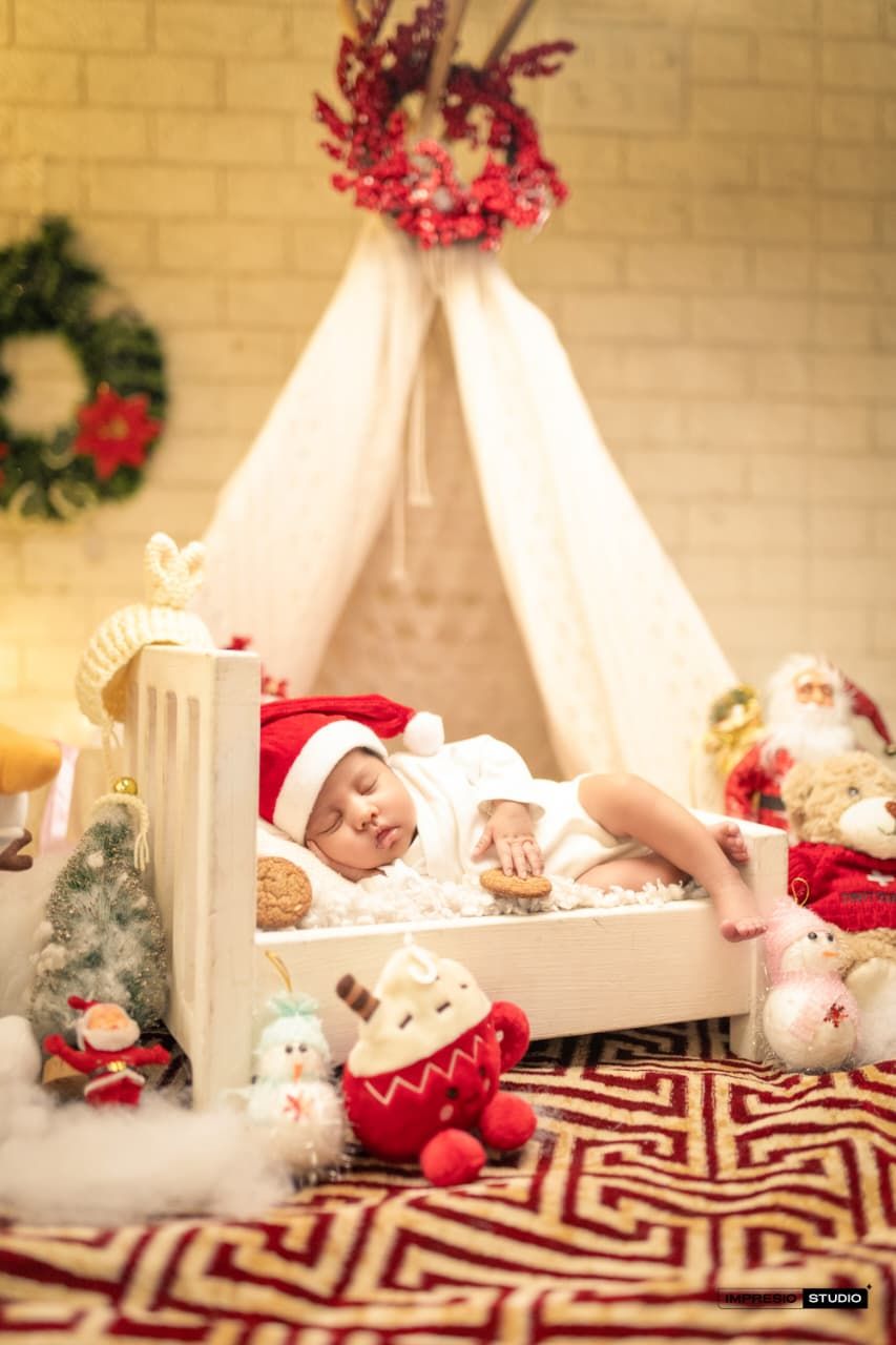 Newborn baby sleeping in white crib wearing Santa hat surrounded by Christmas toys and decorations.