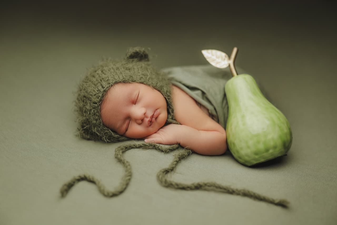 Sleeping newborn baby in green knit hat and wrap posed next to a green pear on a sage background.