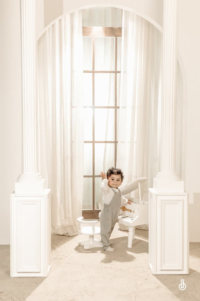 Toddler in gray overalls playing near white columns and arched doorway with sheer curtains.