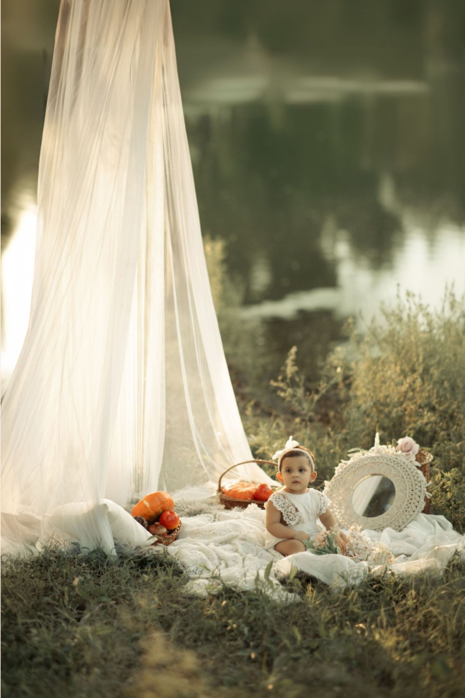 Baby girl in white dress sitting on blanket near lake with sheer canopy, basket of fruit, and straw hat.
