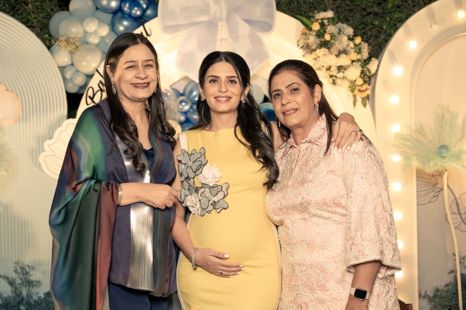 Three women pose together at a baby shower, with the pregnant woman in yellow dress holding her belly, surrounded by blue balloon decor.
