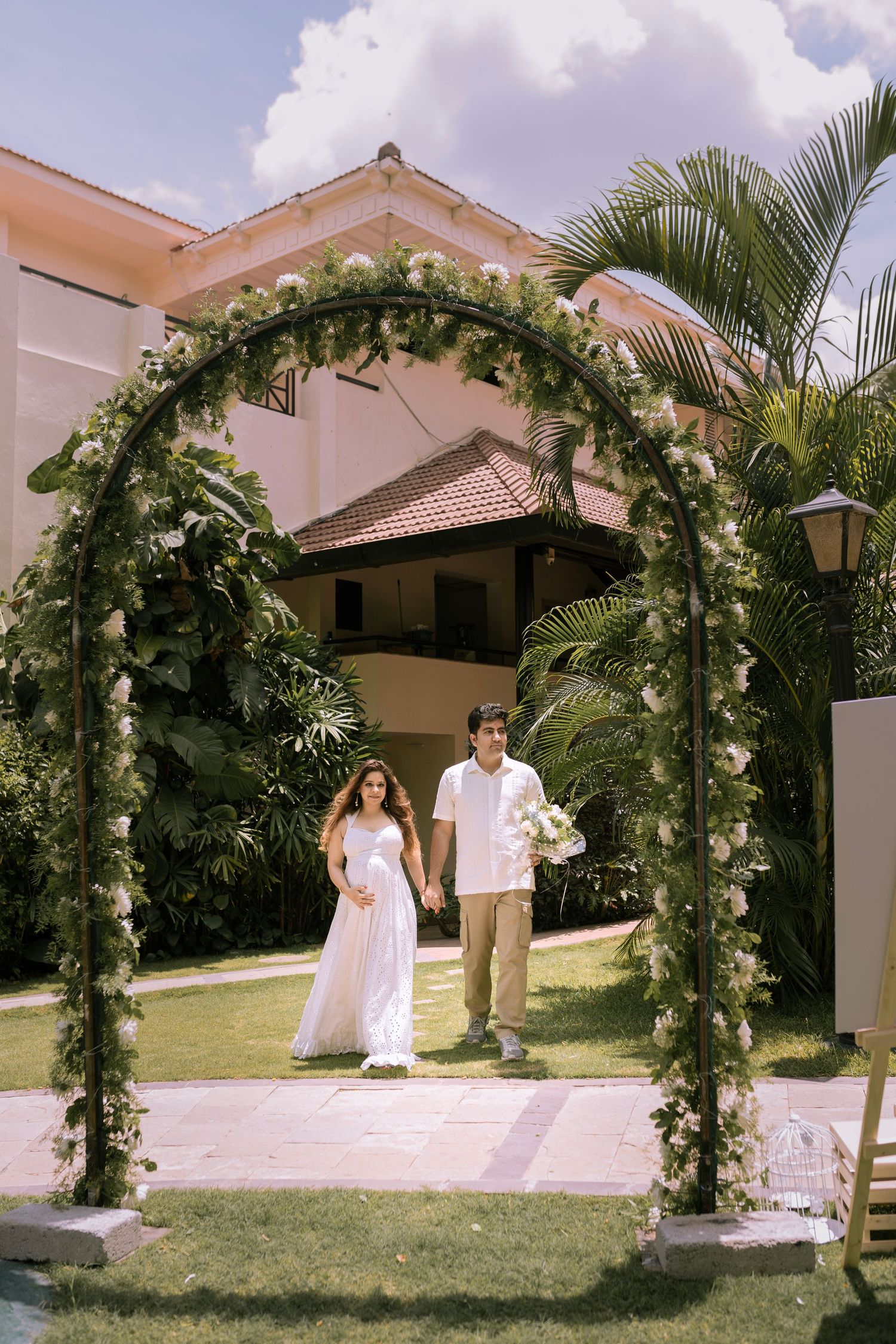 Couple in wedding attire walking under a floral arch with white flowers and greenery at an outdoor tropical venue.