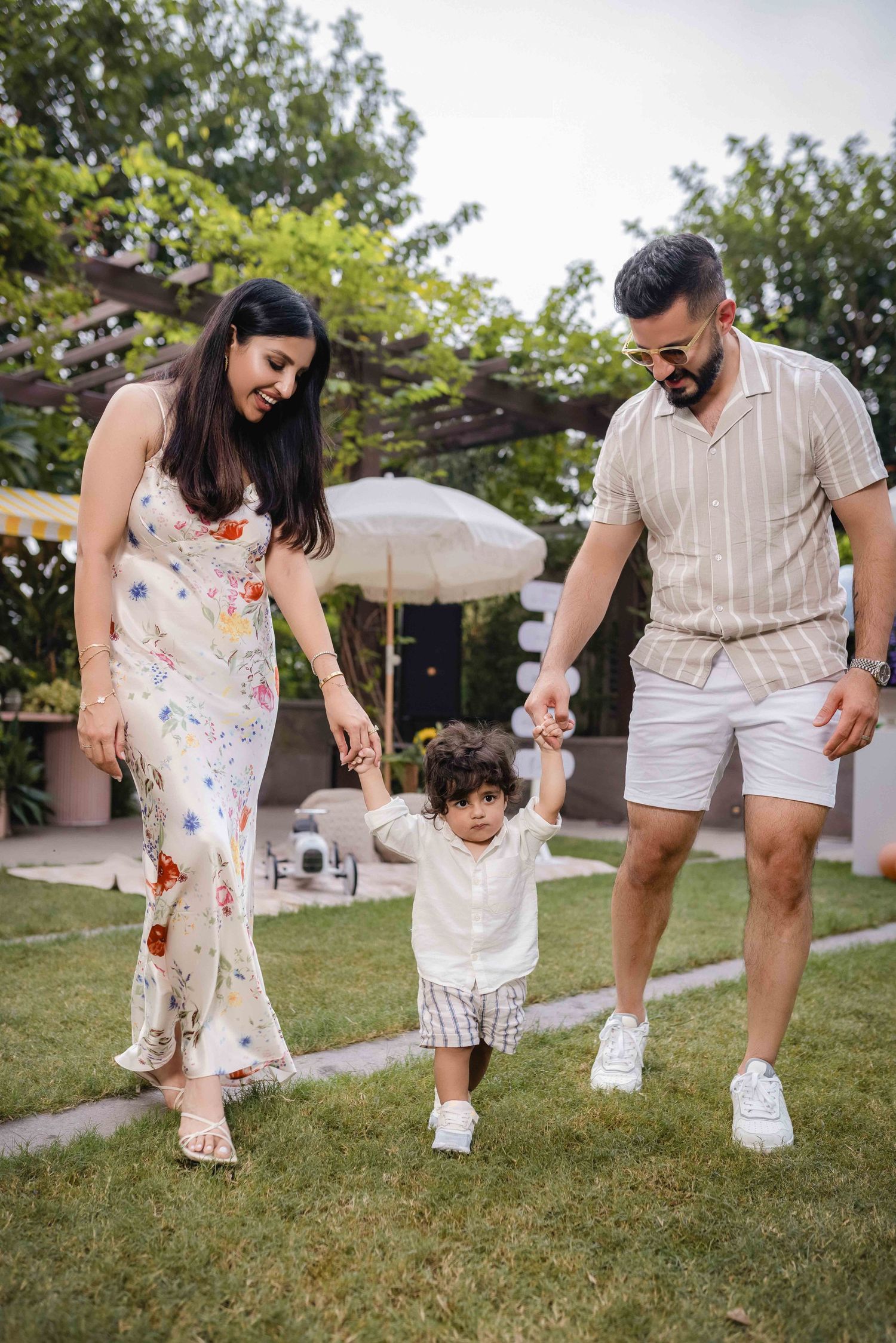 Happy family with parents holding toddler's hands while walking on green lawn in a garden setting.