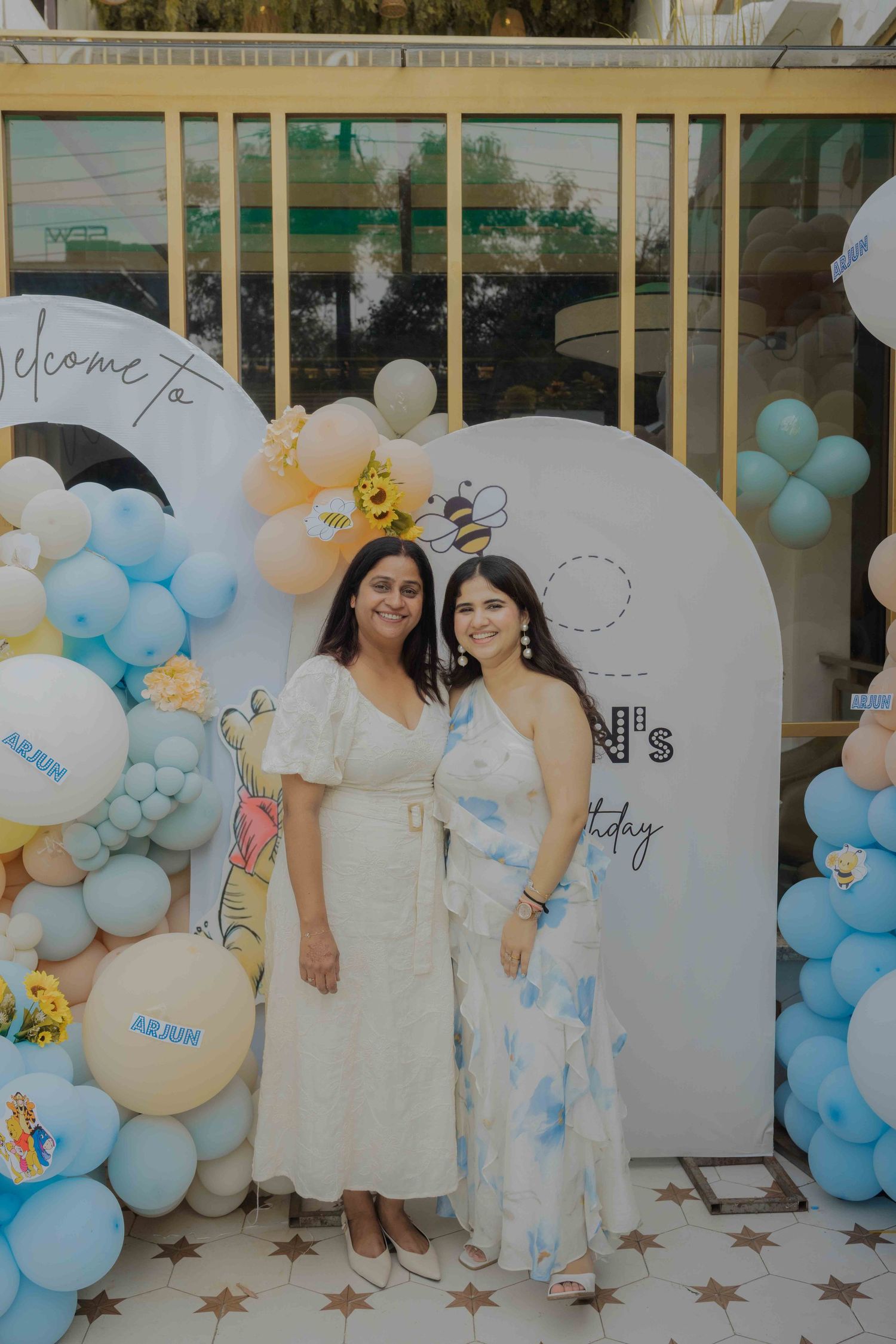 Two women posing together at a birthday party backdrop with blue and yellow balloon decorations.