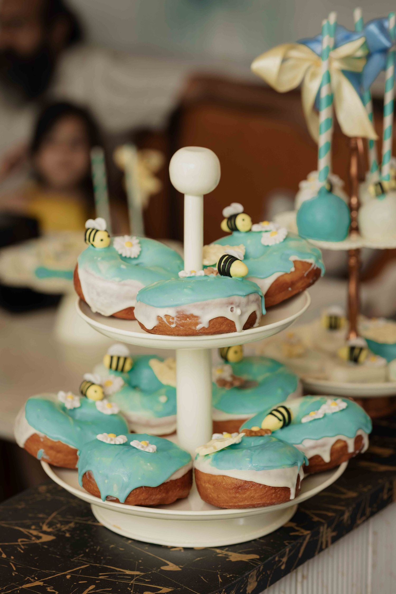 Two-tier stand displaying teal frosted donuts decorated with bee and daisy fondant toppers at a party dessert table.