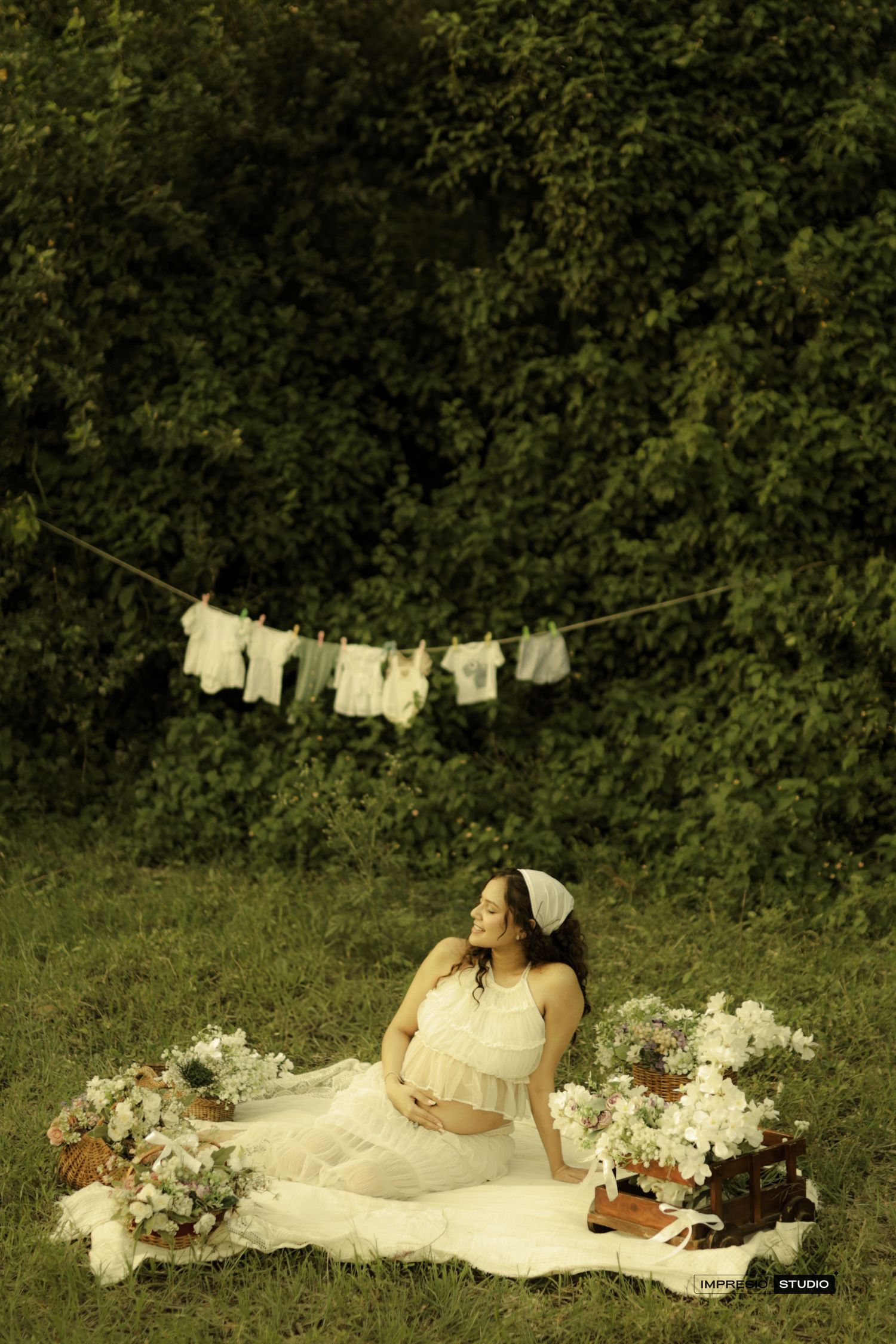 Pregnant woman in white dress sitting on blanket outdoors with flowers and baby clothes hanging on a line.