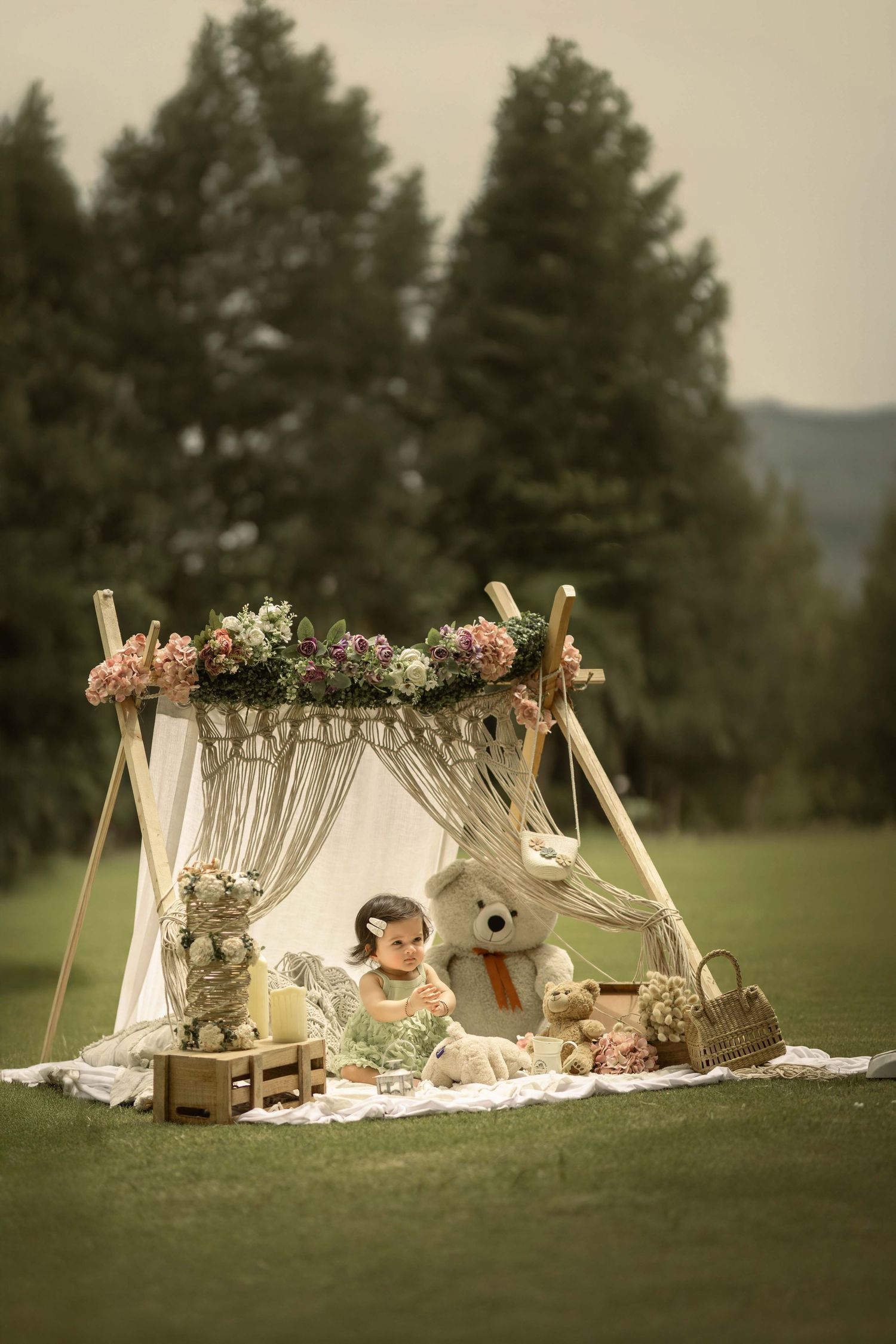A baby sits under a floral boho teepee tent with a teddy bear on a green lawn surrounded by trees.