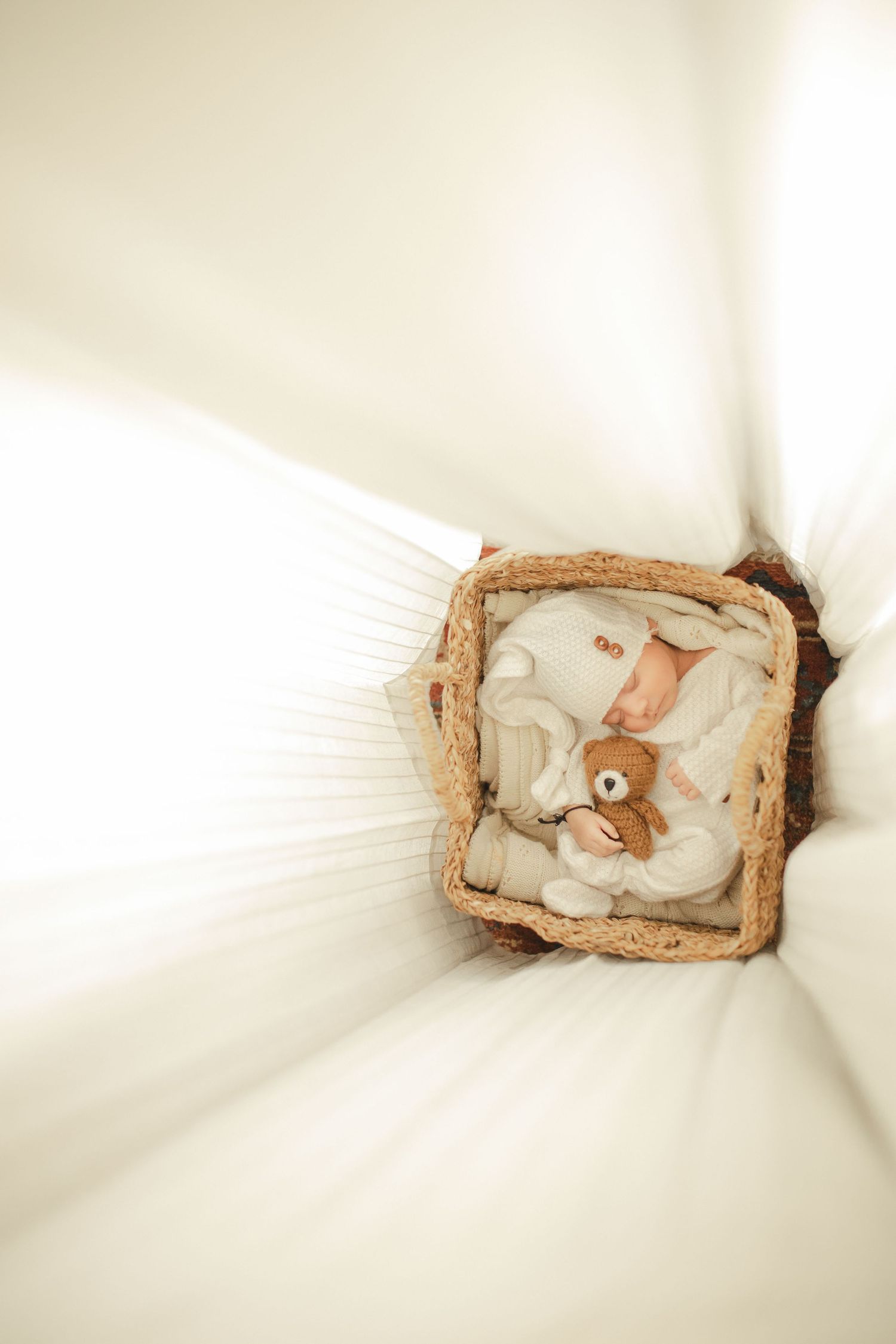 Newborn baby sleeping peacefully in a wicker basket with a teddy bear, dressed in white knit outfit.
