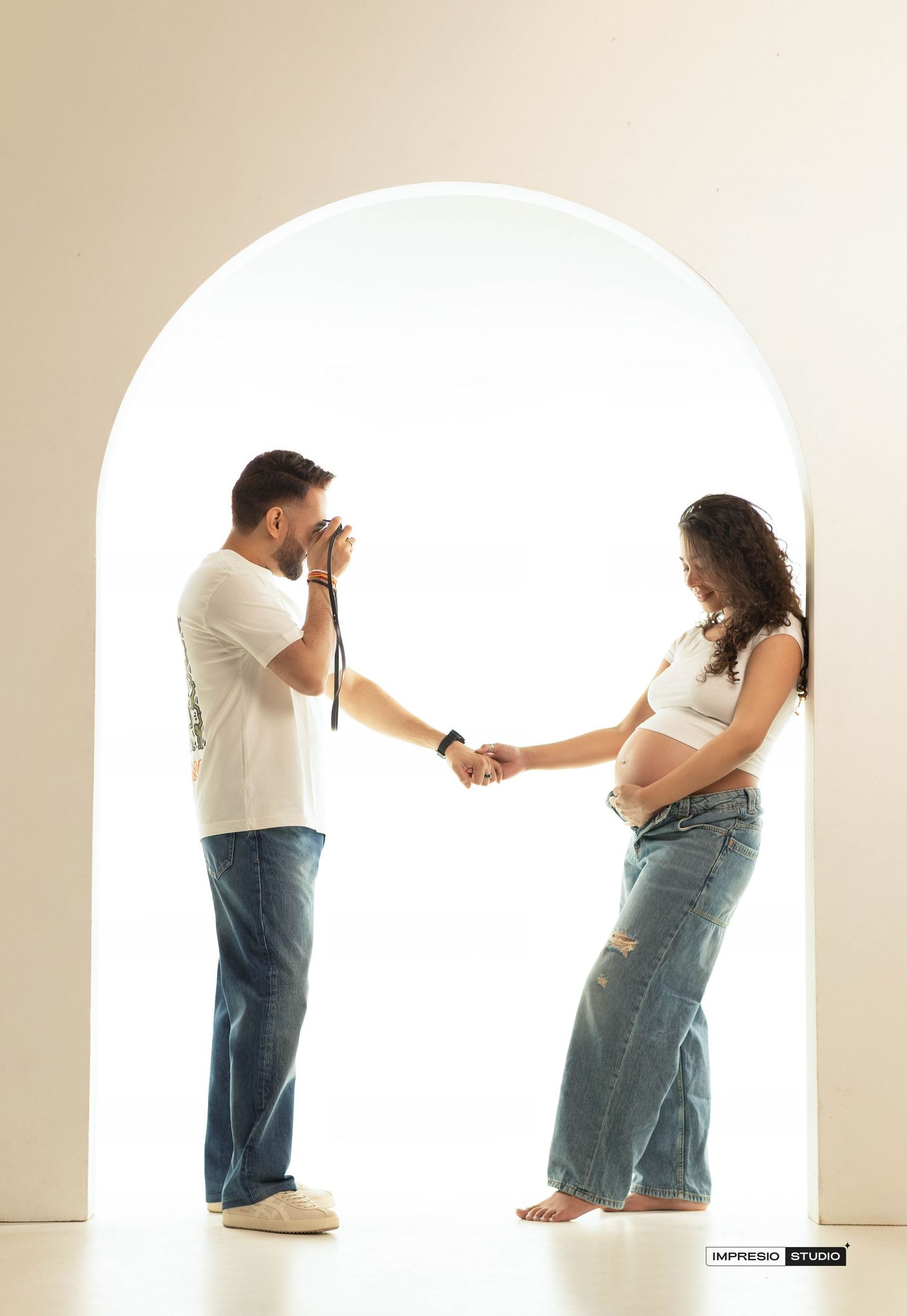 A man photographs his pregnant partner holding hands under a white arch in a bright studio maternity shoot.
