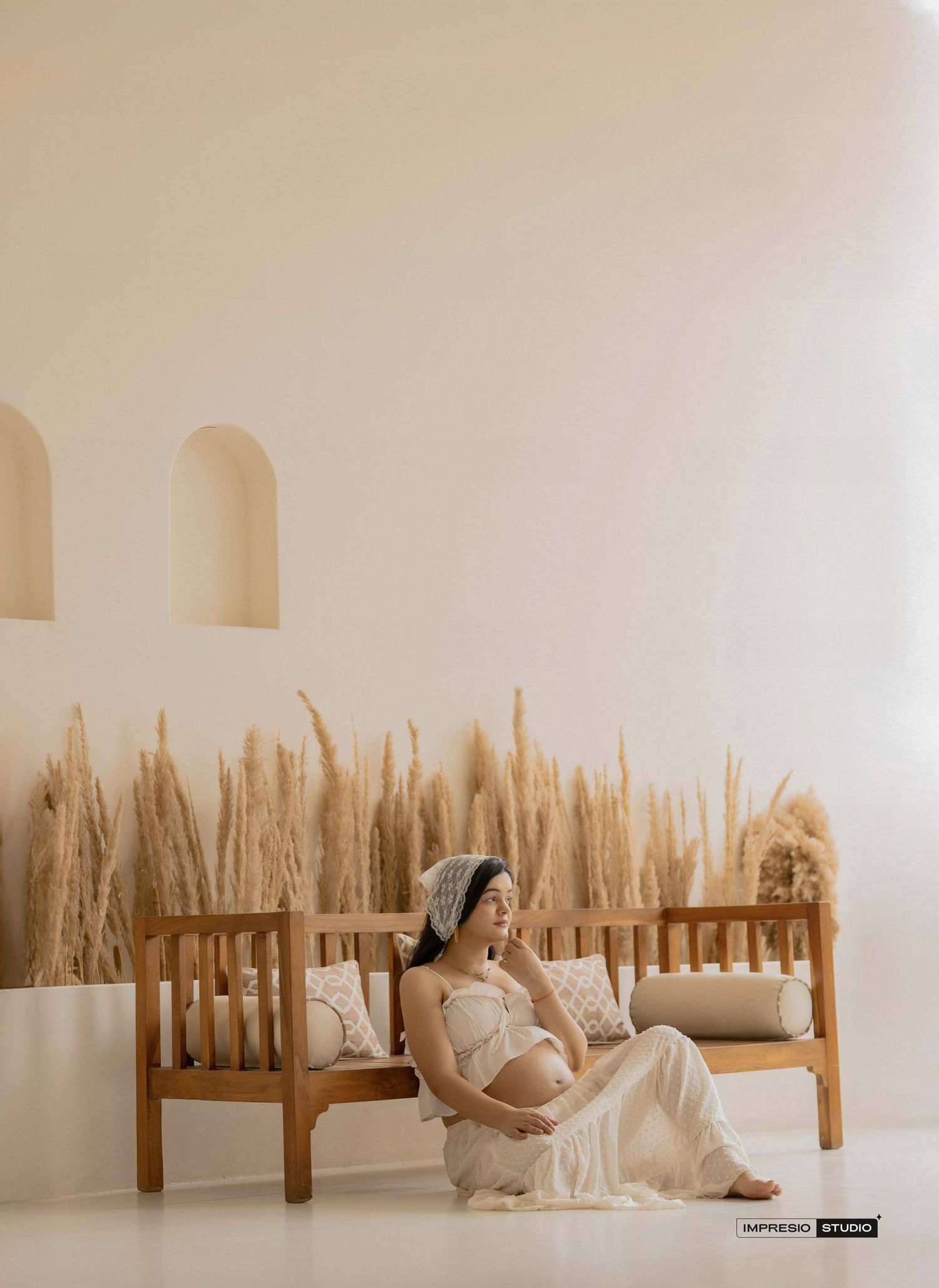 Pregnant woman in white dress sitting on wooden bench with pampas grass decor in minimalist beige studio.