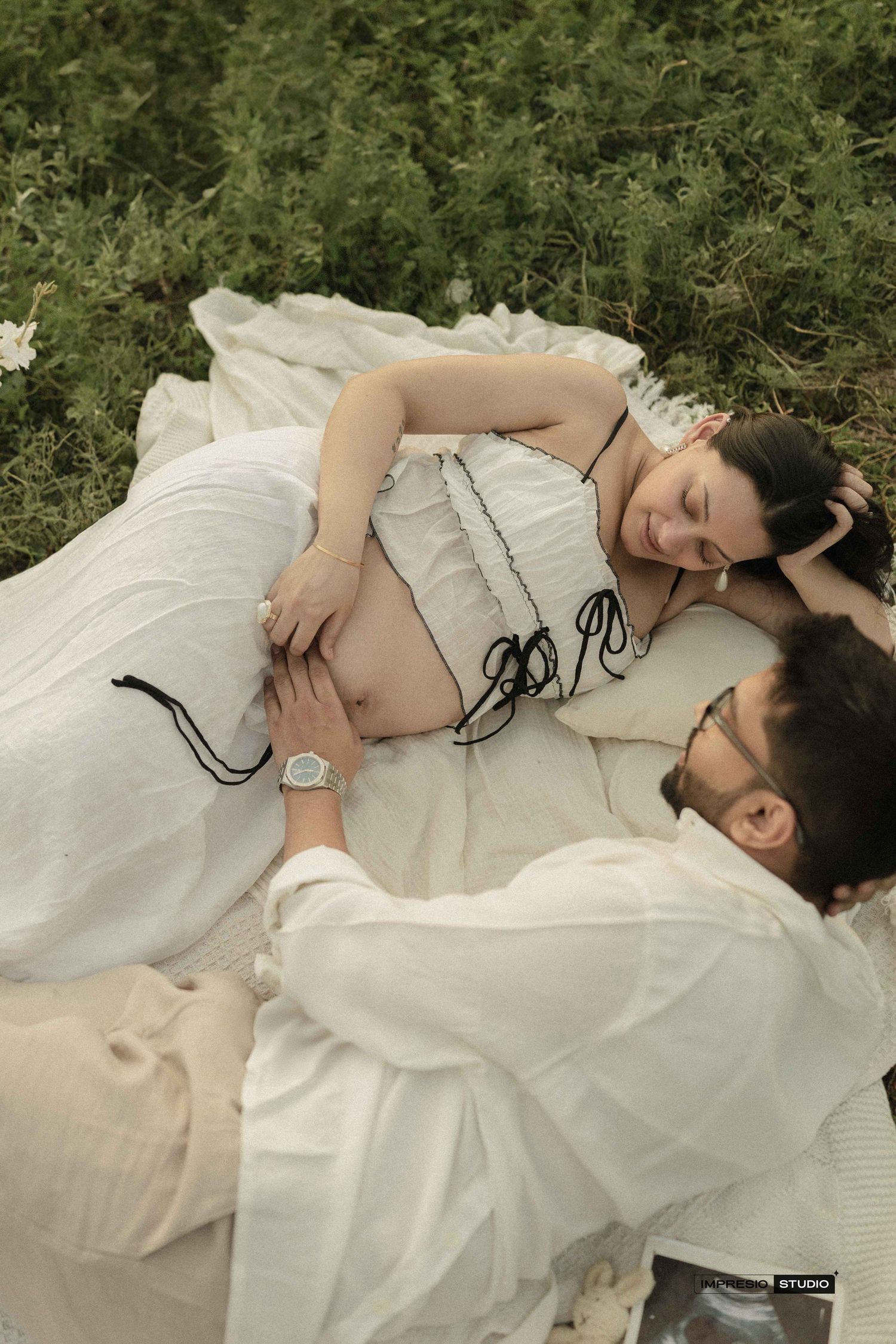 Pregnant woman in white dress lying on grass with partner tenderly touching her baby bump during maternity photoshoot.
