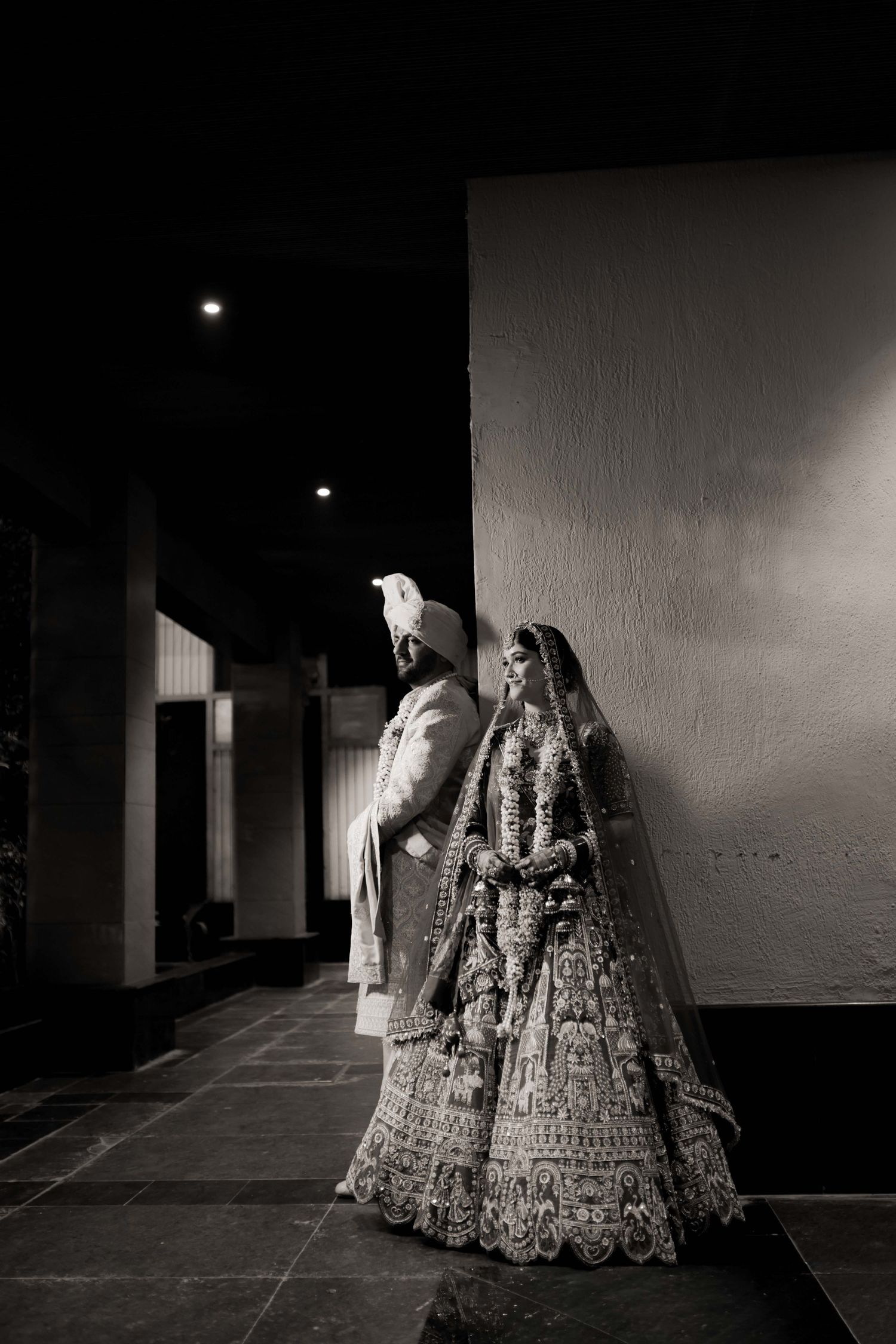 Black and white photo of an Indian bride and groom in traditional wedding attire posing elegantly against a wall at night.