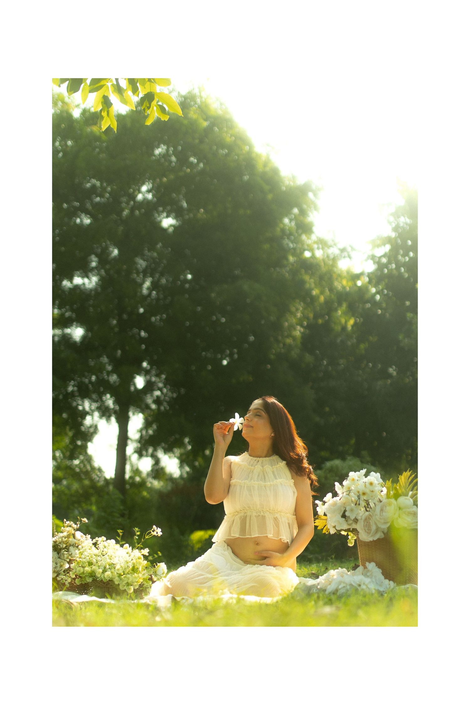 A person in a white dress lies on grass in a sunlit garden setting with trees and flowers in the background.