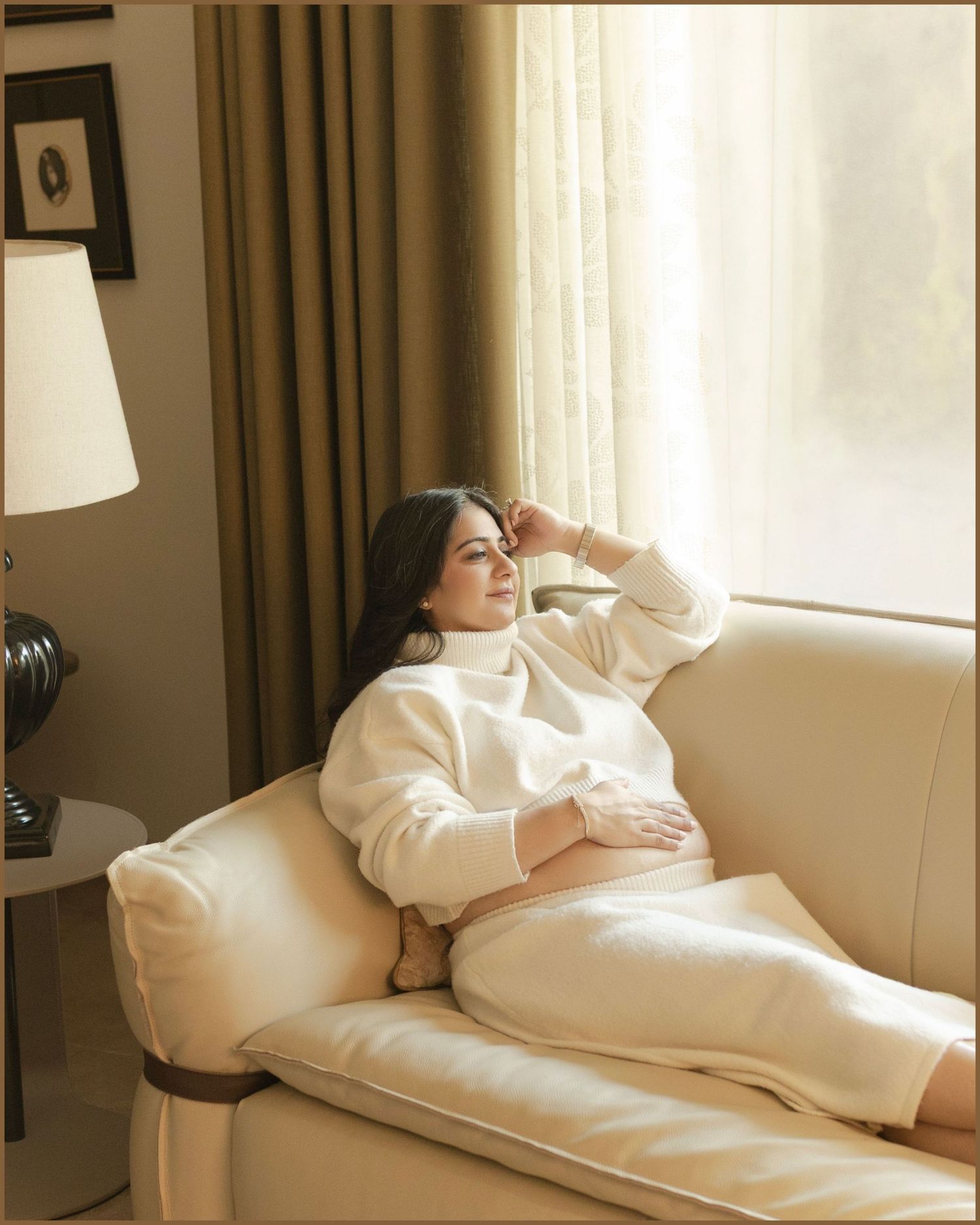 A woman in a white turtleneck sweater and skirt relaxes on a cream sofa by a sunlit window.