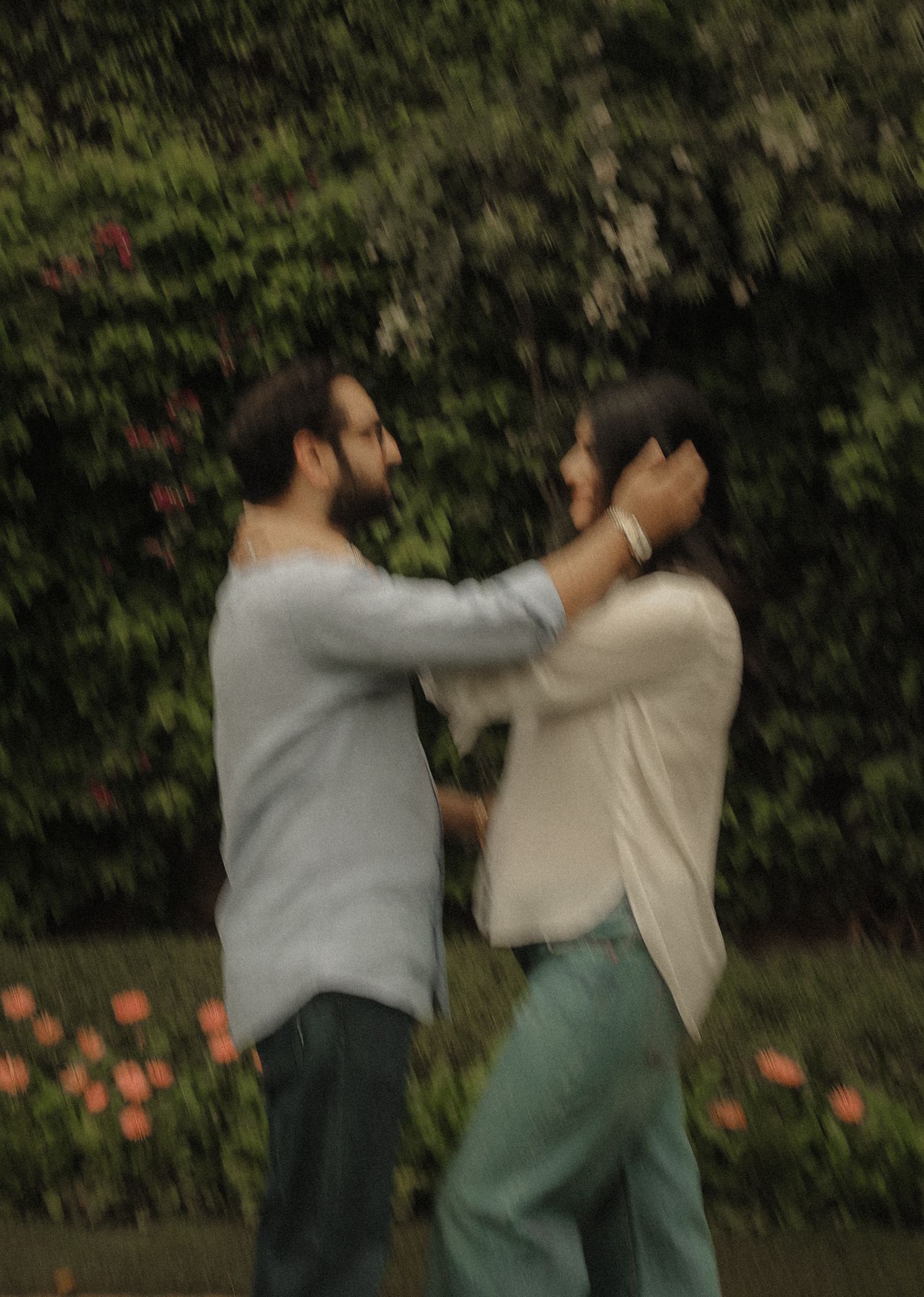 A couple shares a tender moment in a lush garden, the man gently touching the woman's face near blooming flowers.