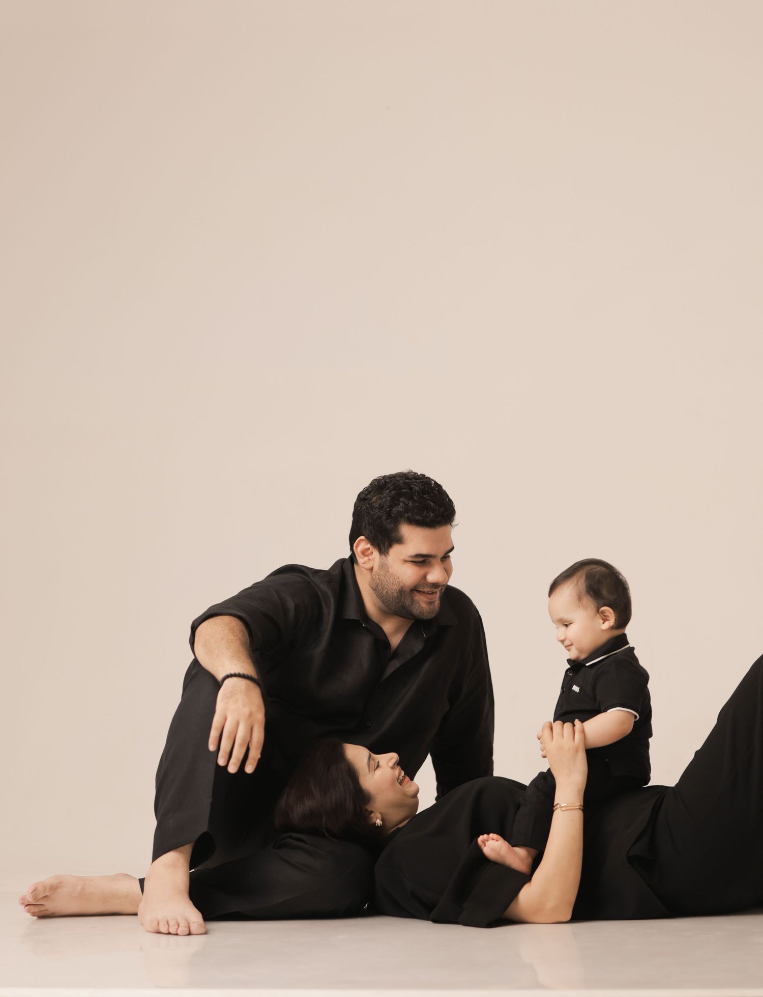 Family in matching black outfits, parents smiling at baby sitting up, studio portrait on neutral background.