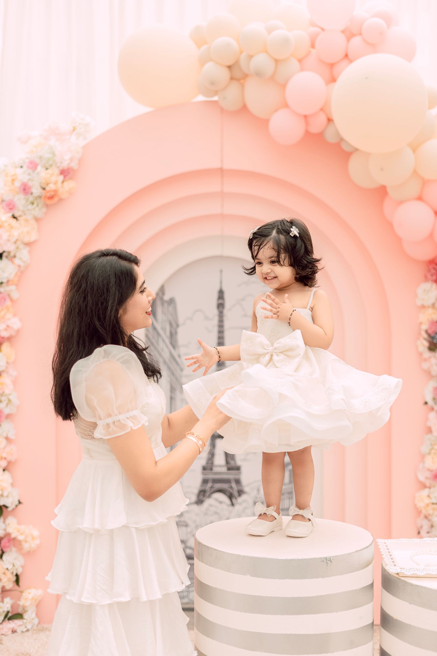 A mother in white dress lifts smiling toddler girl in white tutu at pink balloon arch birthday party.