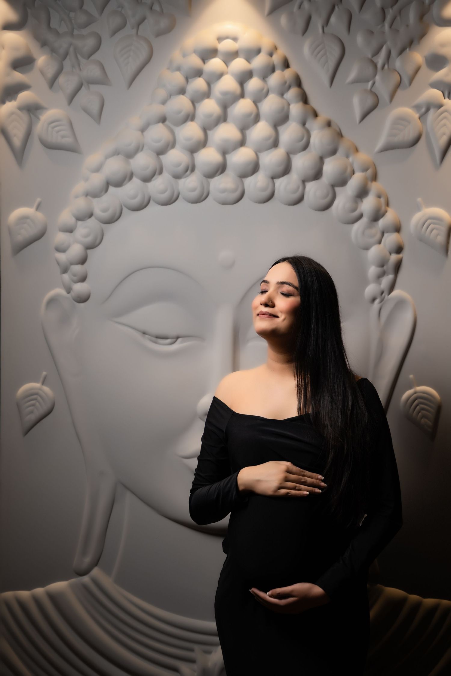 Pregnant woman in black off-shoulder dress poses serenely in front of a white Buddha wall sculpture.