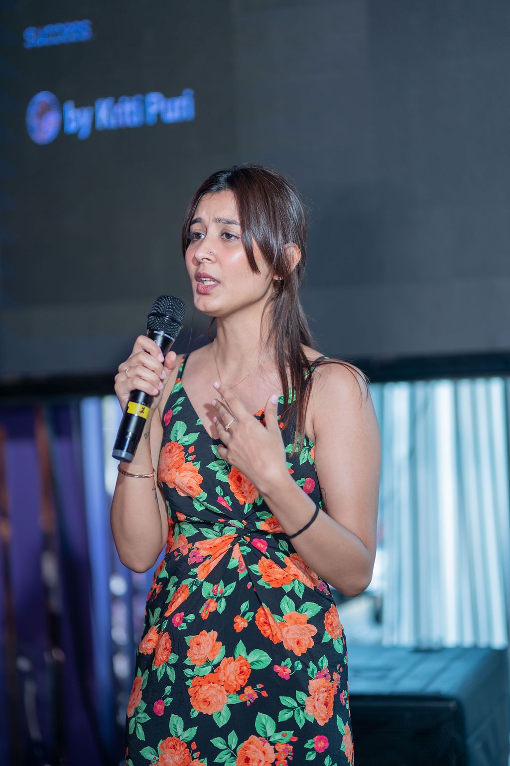 A performer in a floral print dress sings into a microphone on stage with blue lighting in the background.