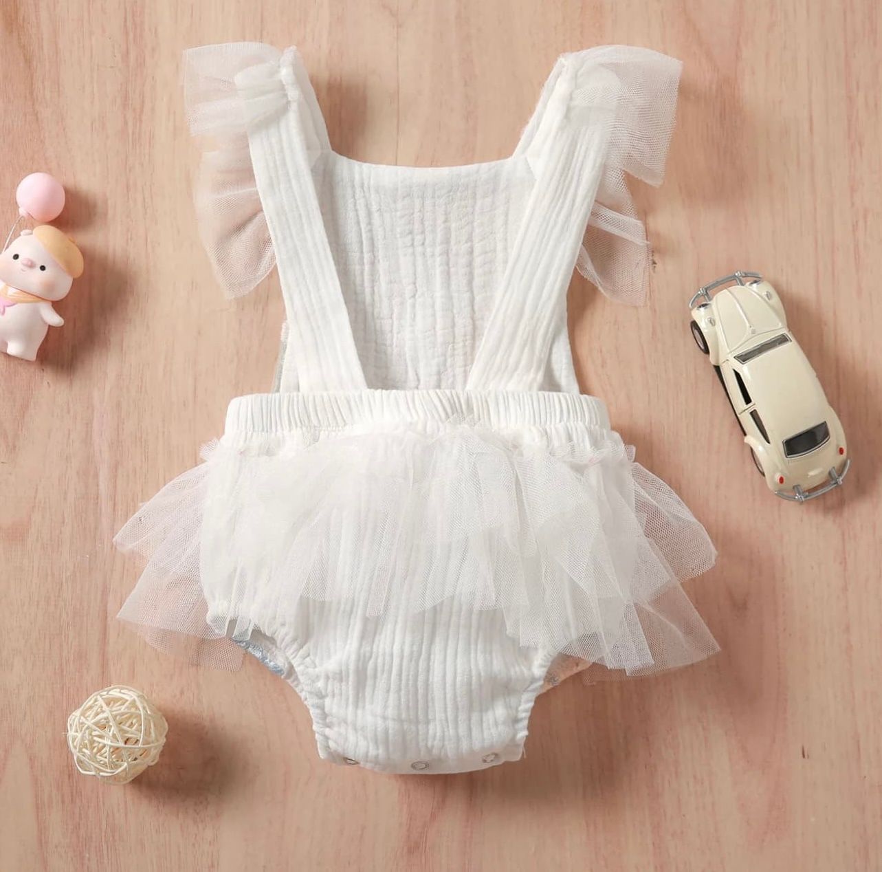 White ruffled tulle romper with matching accessories and small toys arranged on a wooden background.