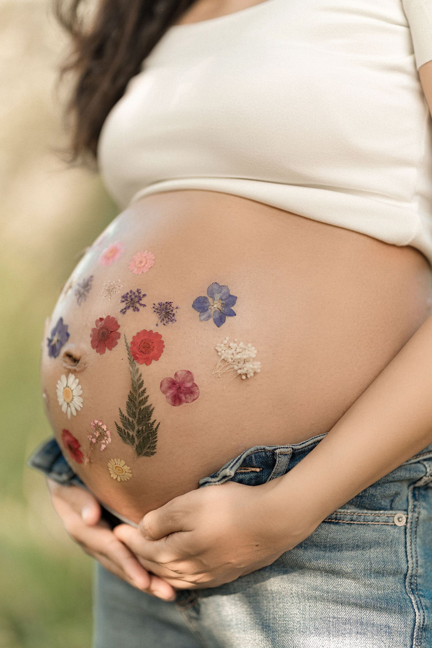 A pregnant belly adorned with delicate pressed flowers in shades of purple, red, and white against blue denim jeans.