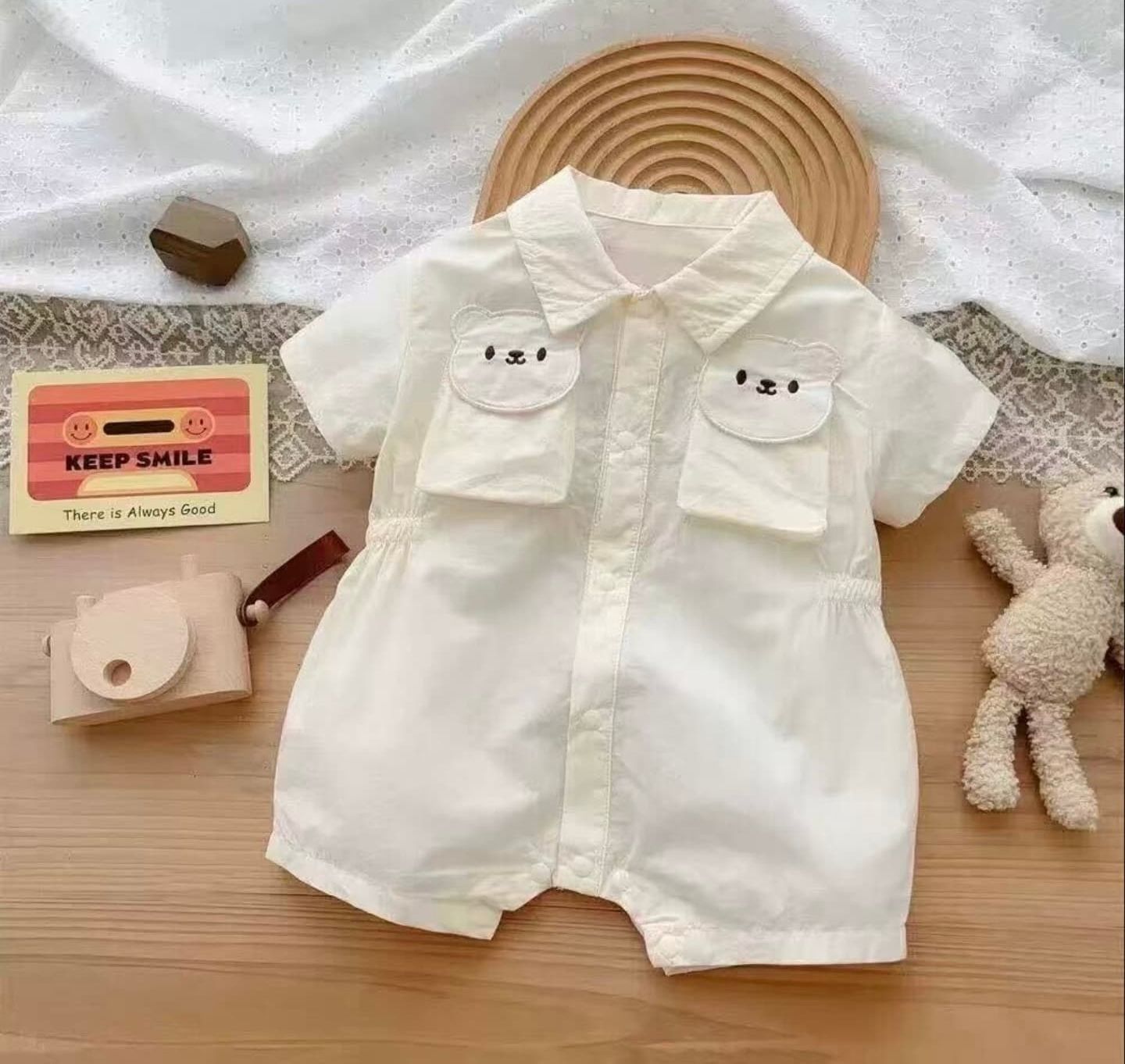 Adorable white baby romper with cute bear pocket designs displayed with plush toy and wooden accessories on table.