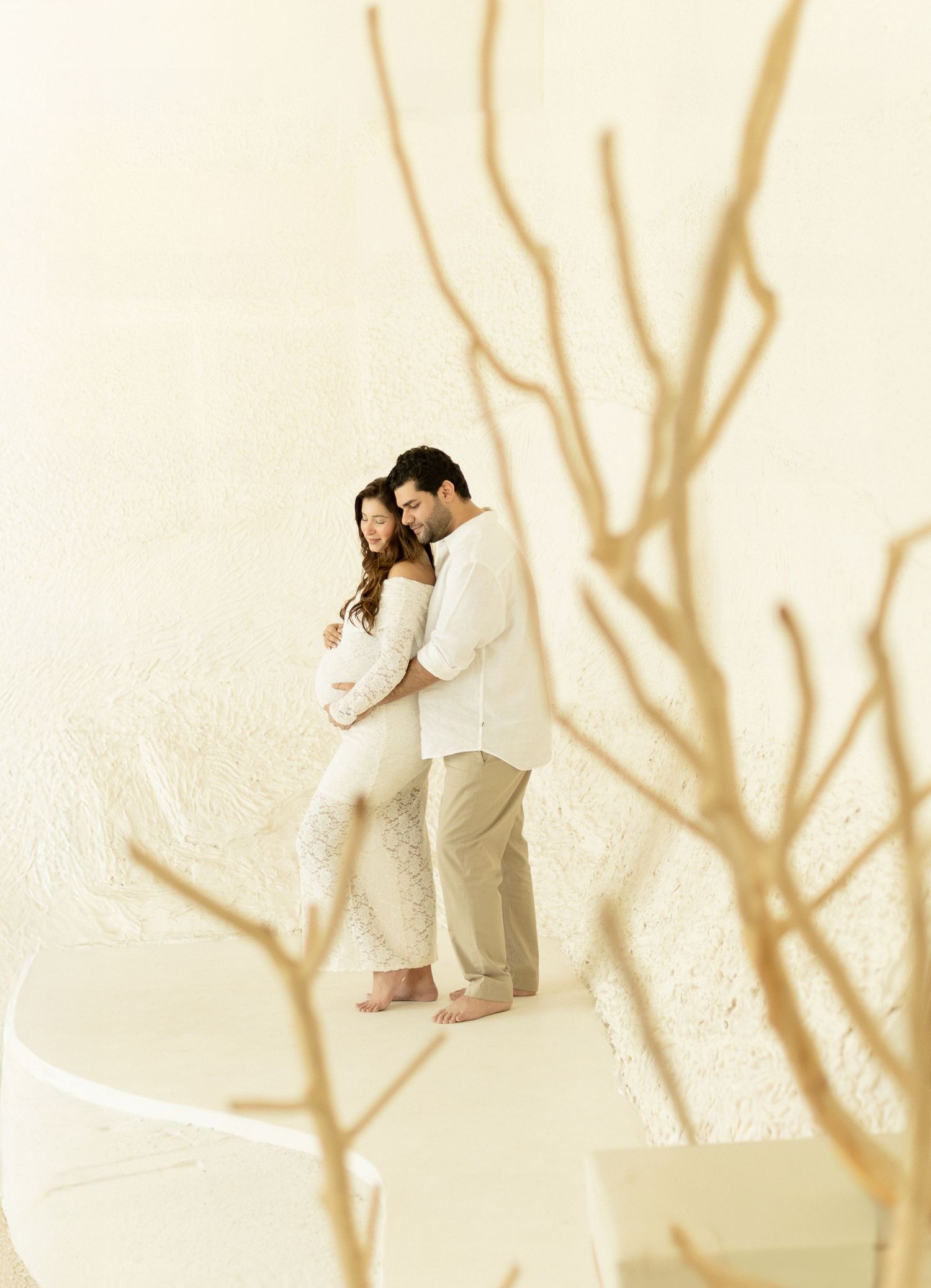 A couple in white outfits embraces and kisses against a beige background with bare tree branches.