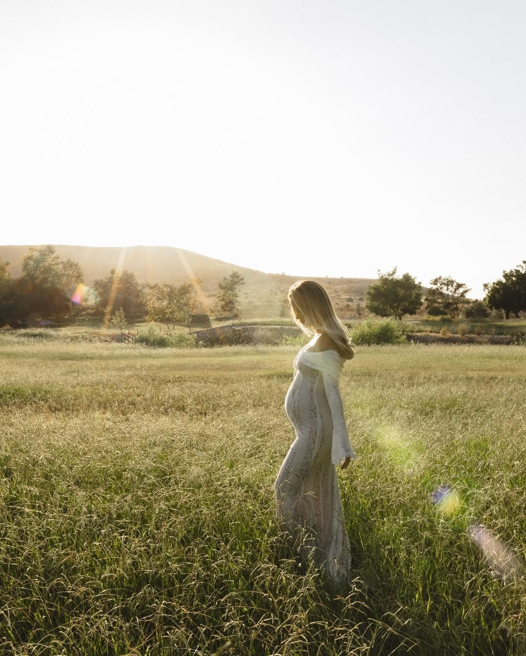 Sun-drenched portrait of expecting mother in fitted lace dress standing in wild meadow.
