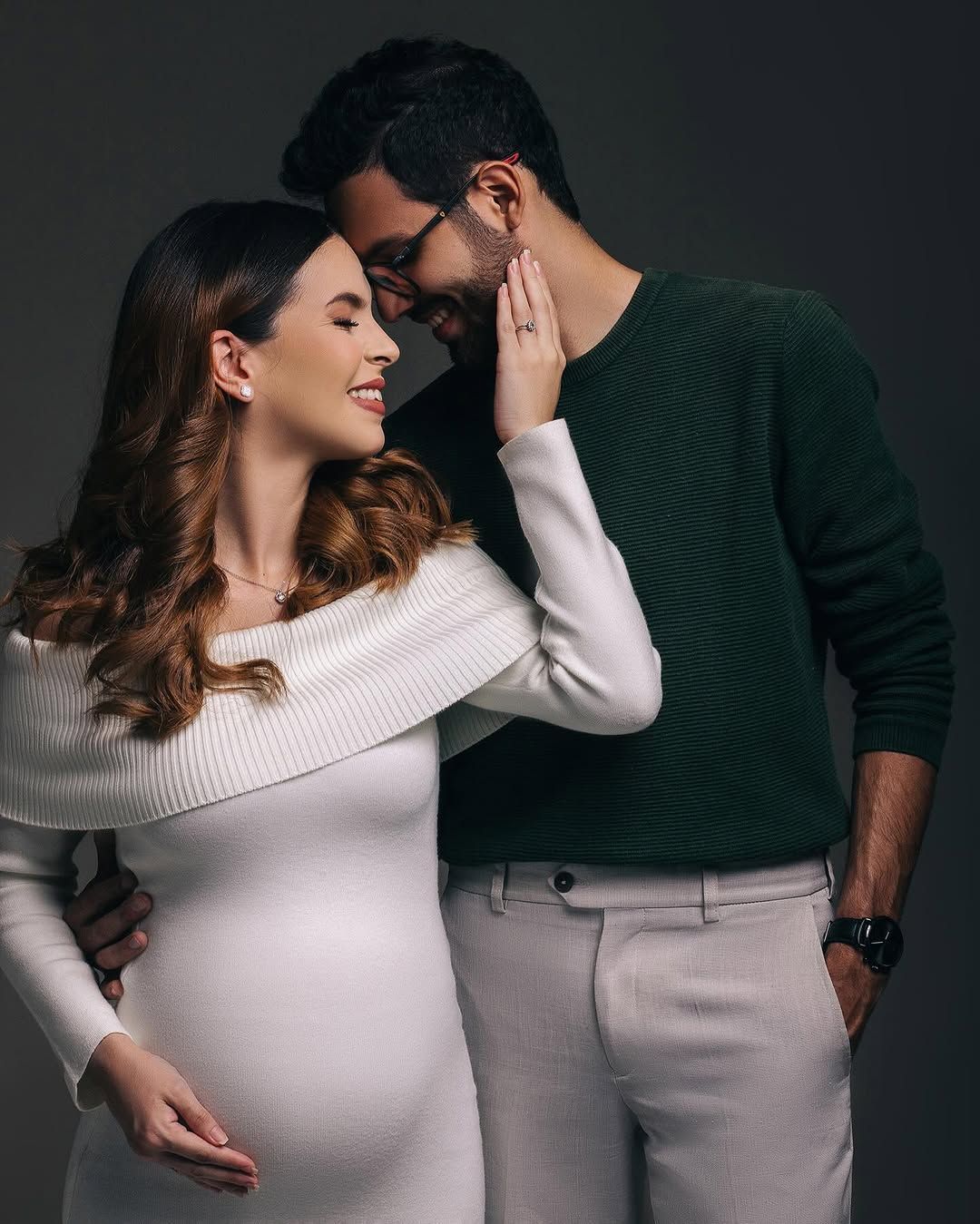 Romantic maternity portrait of couple in white dress and green sweater embracing against neutral background.