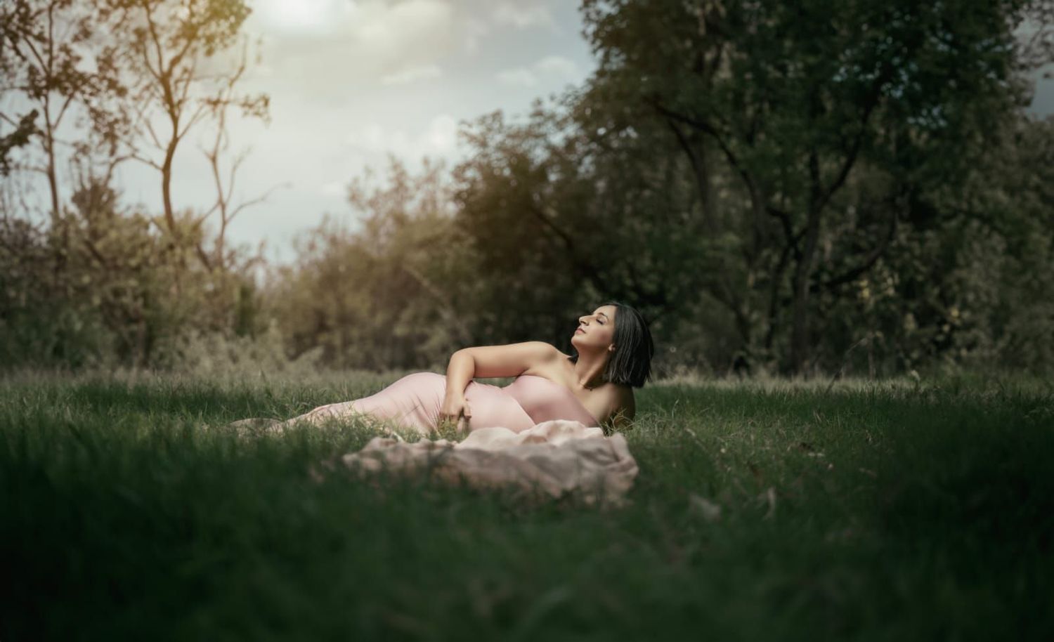 Ethereal maternity photo of woman in pink dress reclining in sunlit meadow.