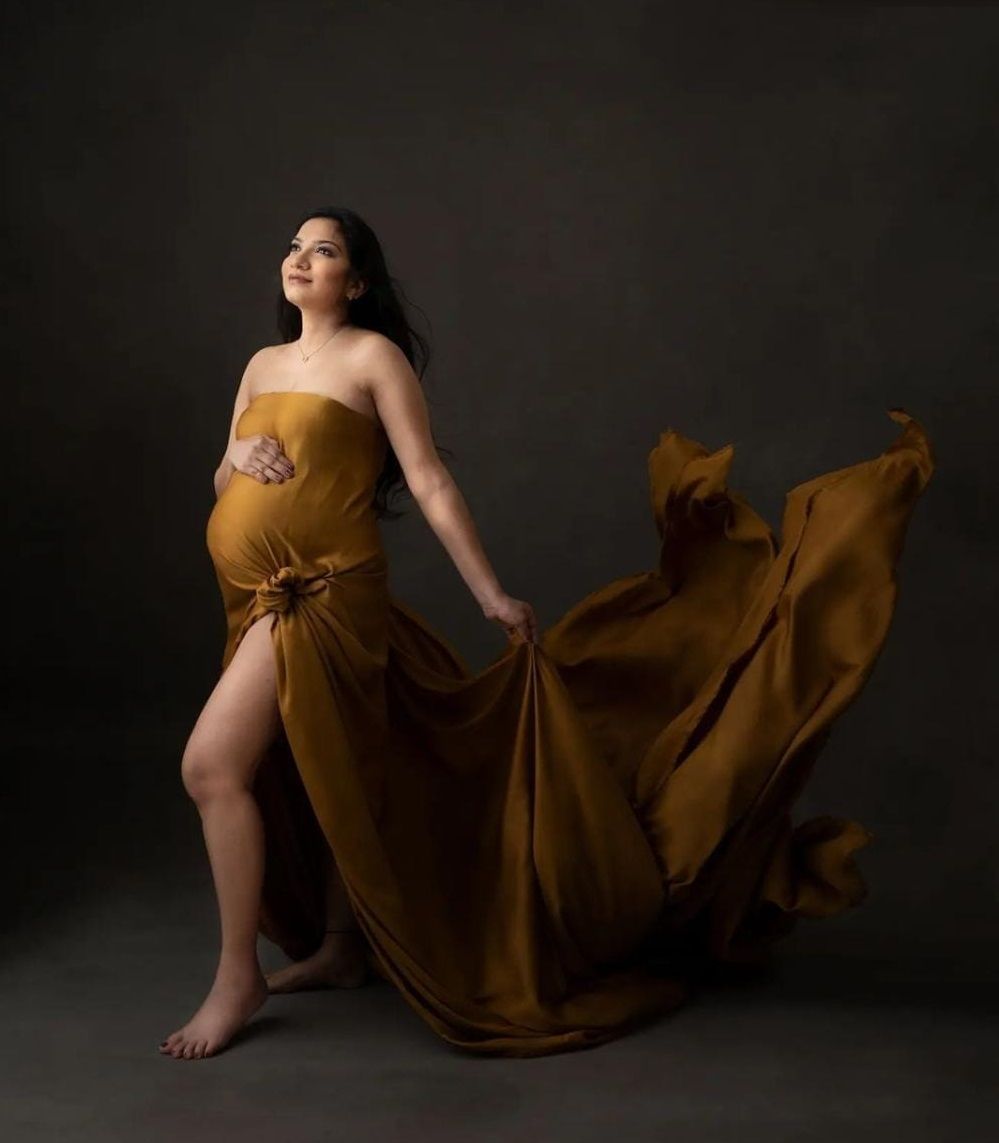 Artistic maternity portraits in flowing mustard dress against dark backdrop.