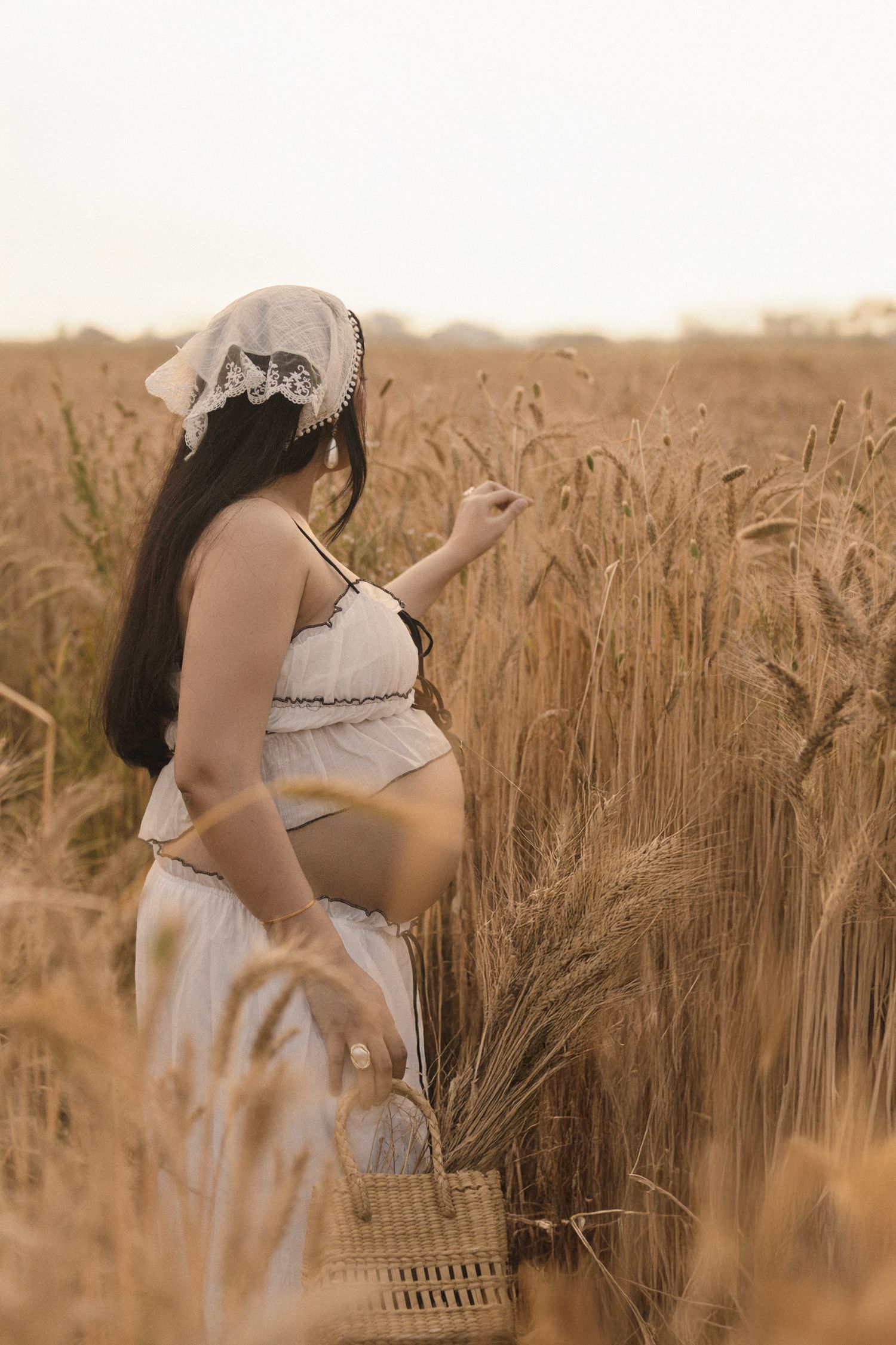 Pregnant woman in white dress and lace headband touching wheat stalks in golden field, holding wicker basket.