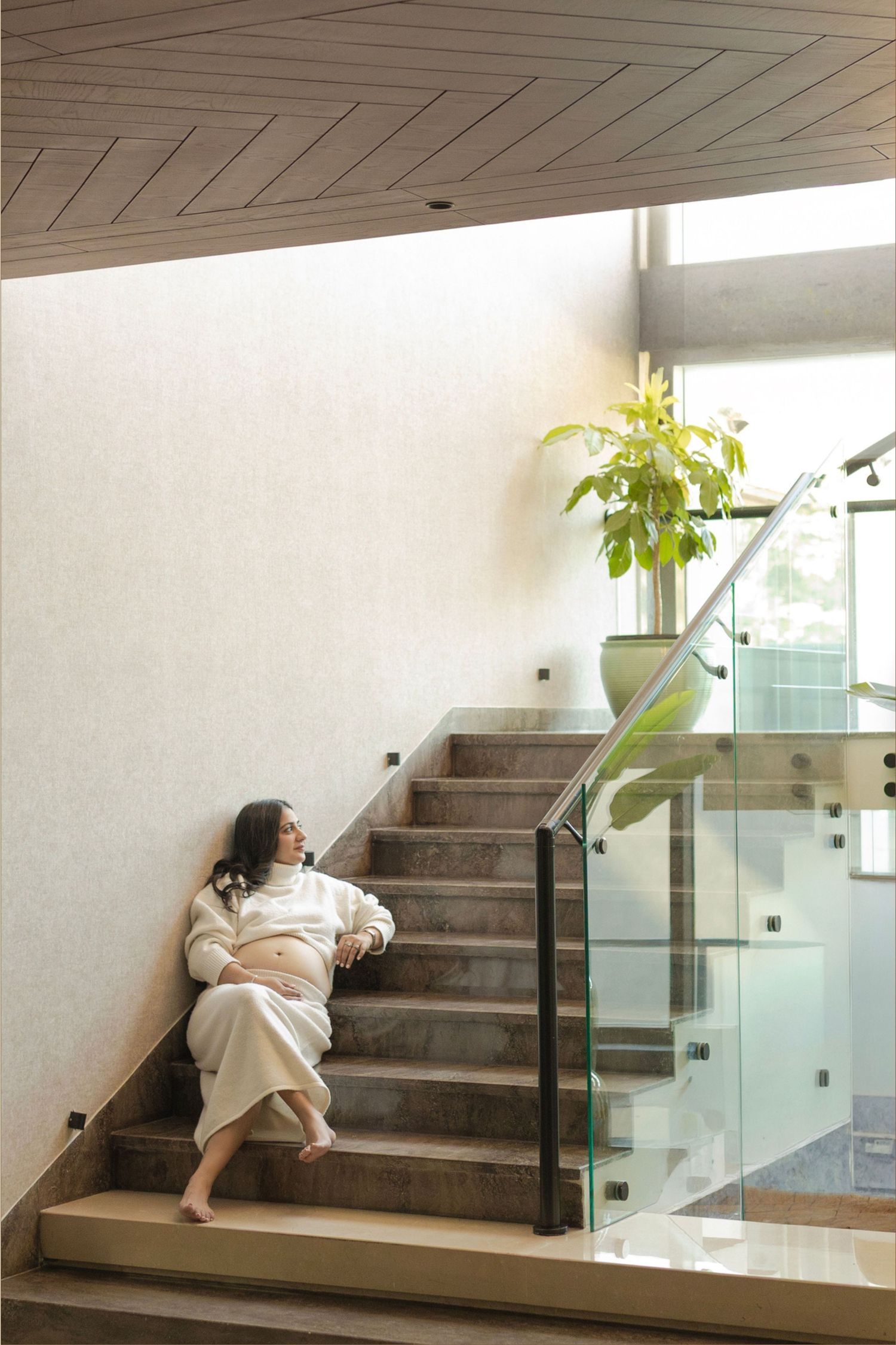 Pregnant woman in white robe sitting relaxed on modern staircase with glass railing and indoor plant.