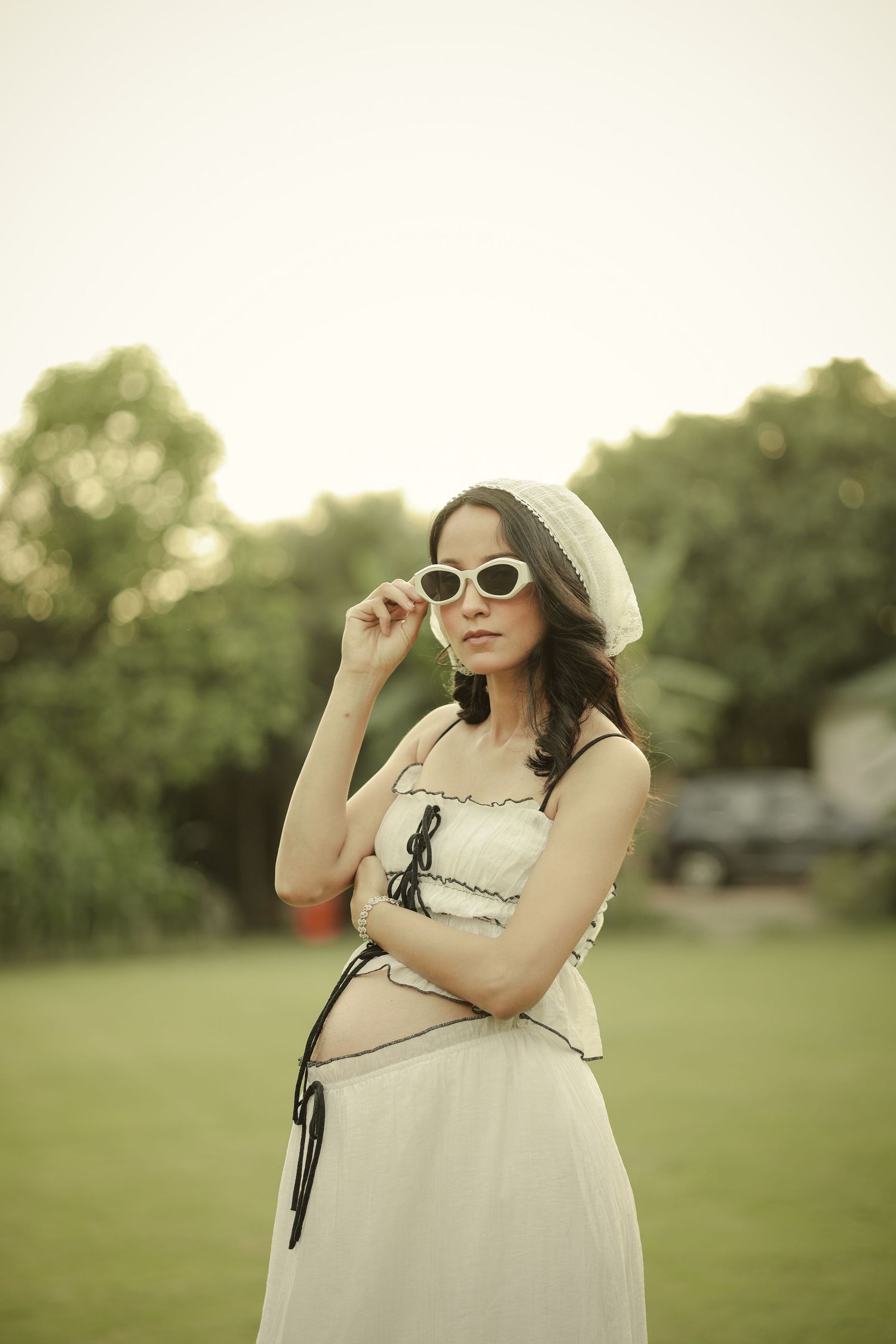 Pregnant woman in white beret and sunglasses posing outdoors in a park wearing a stylish white crop top and skirt.