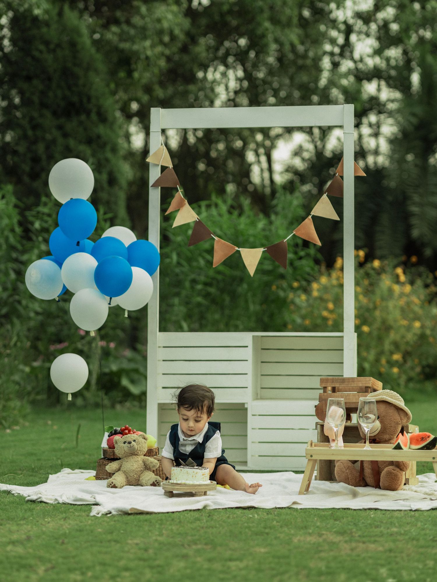 Toddler boy sits outdoors at a birthday setup with teddy bears, balloons, bunting, and watermelon on a white blanket.