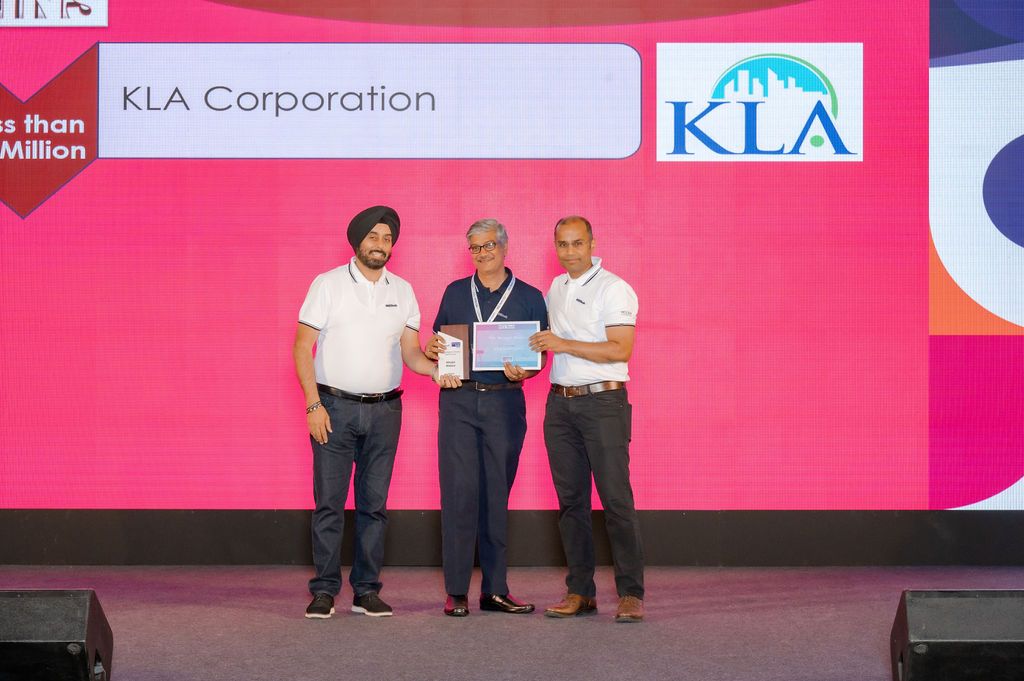 Three people stand on a red-lit stage with a KLA Corporation logo display behind them at a corporate event.