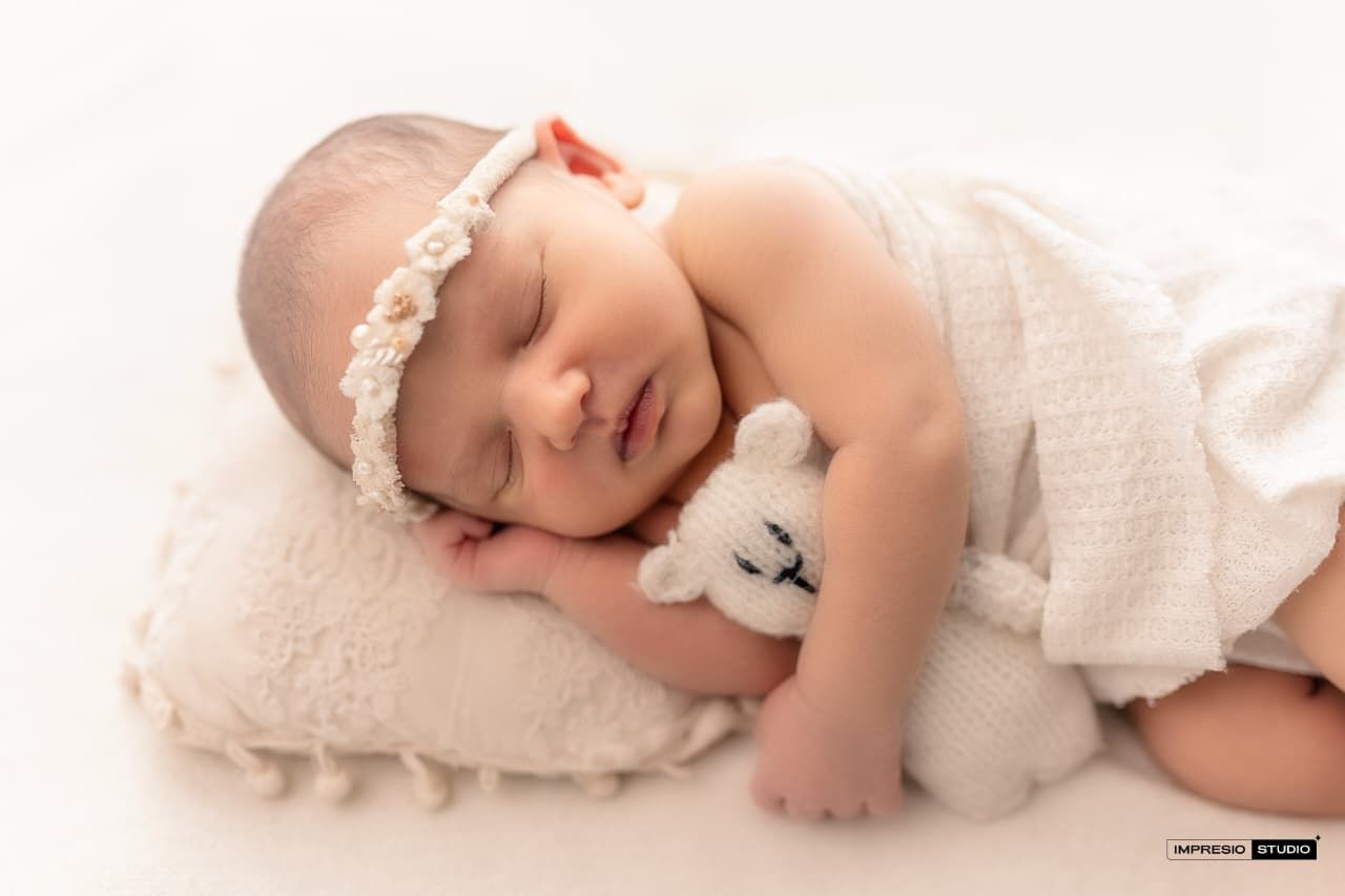 Sleeping newborn baby girl with floral headband cuddles a tiny bear toy on a soft cream pillow.