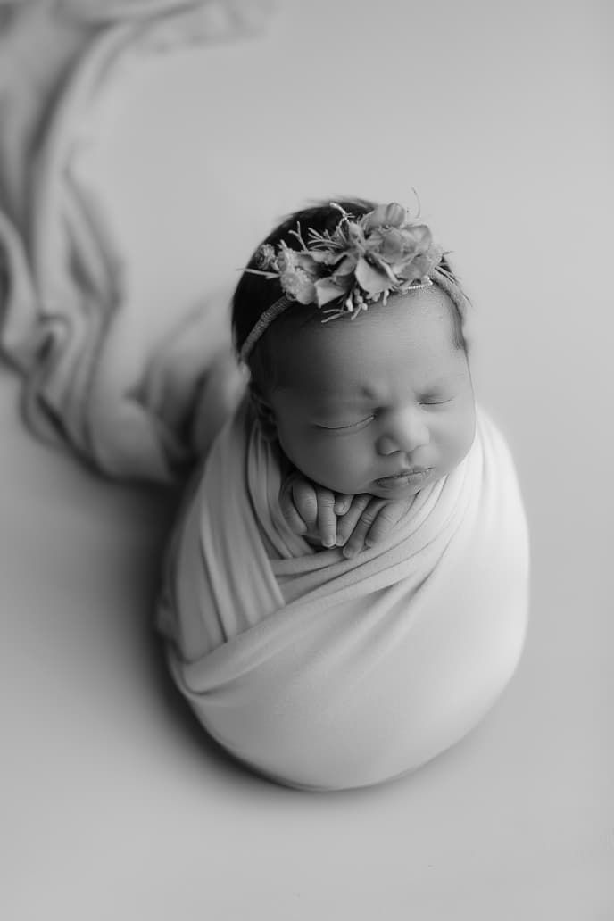 Black and white photo of a sleeping newborn baby girl swaddled in cloth, wearing a floral headband.
