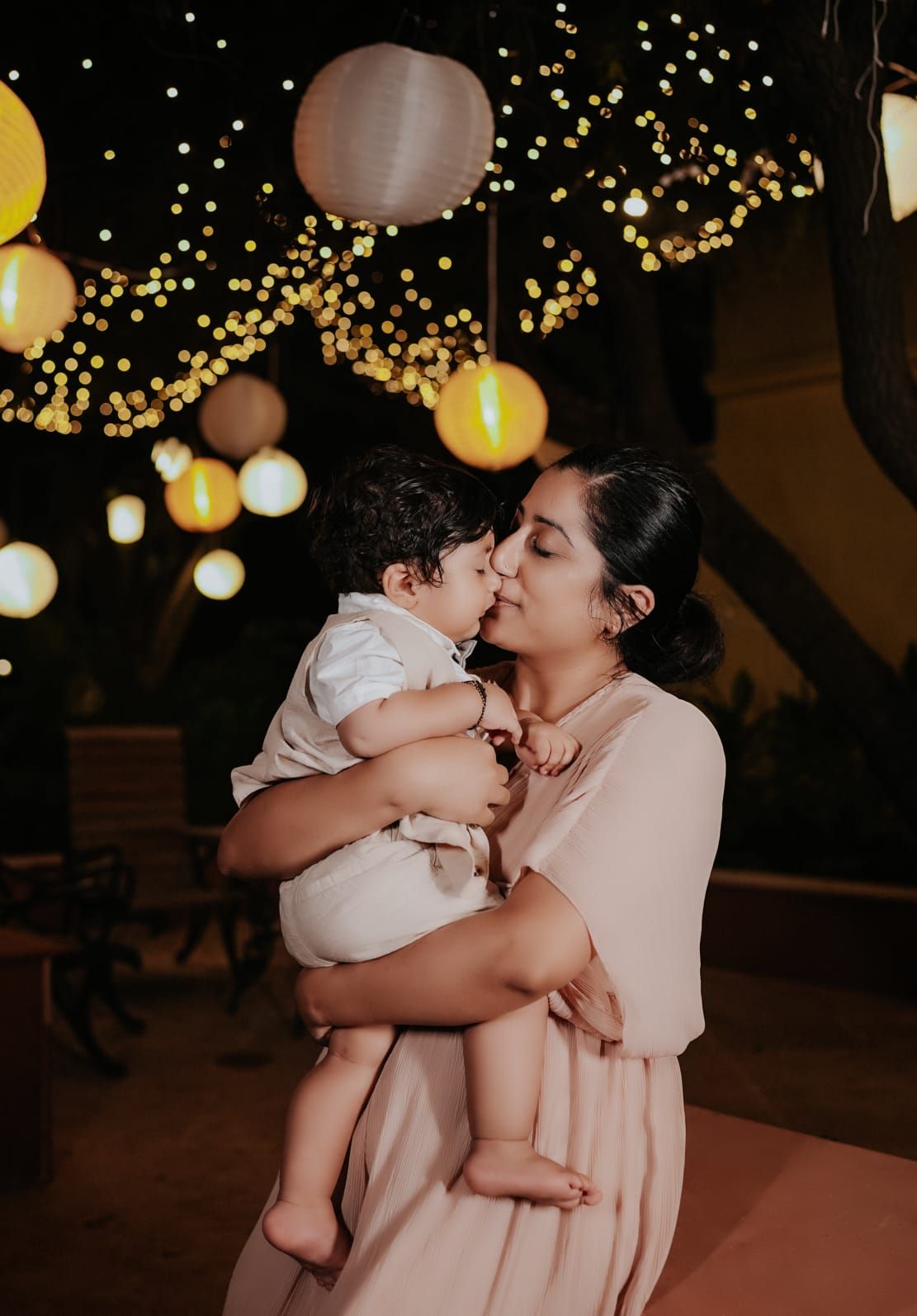 A woman tenderly kisses her baby boy under glowing paper lanterns and fairy lights at a nighttime celebration.