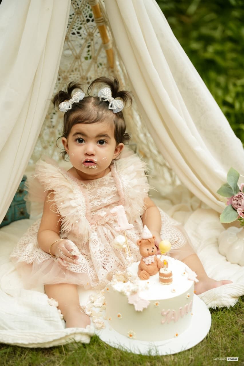 Toddler girl in pink feather dress sits beside a bear-topped birthday cake inside a boho teepee outdoors.