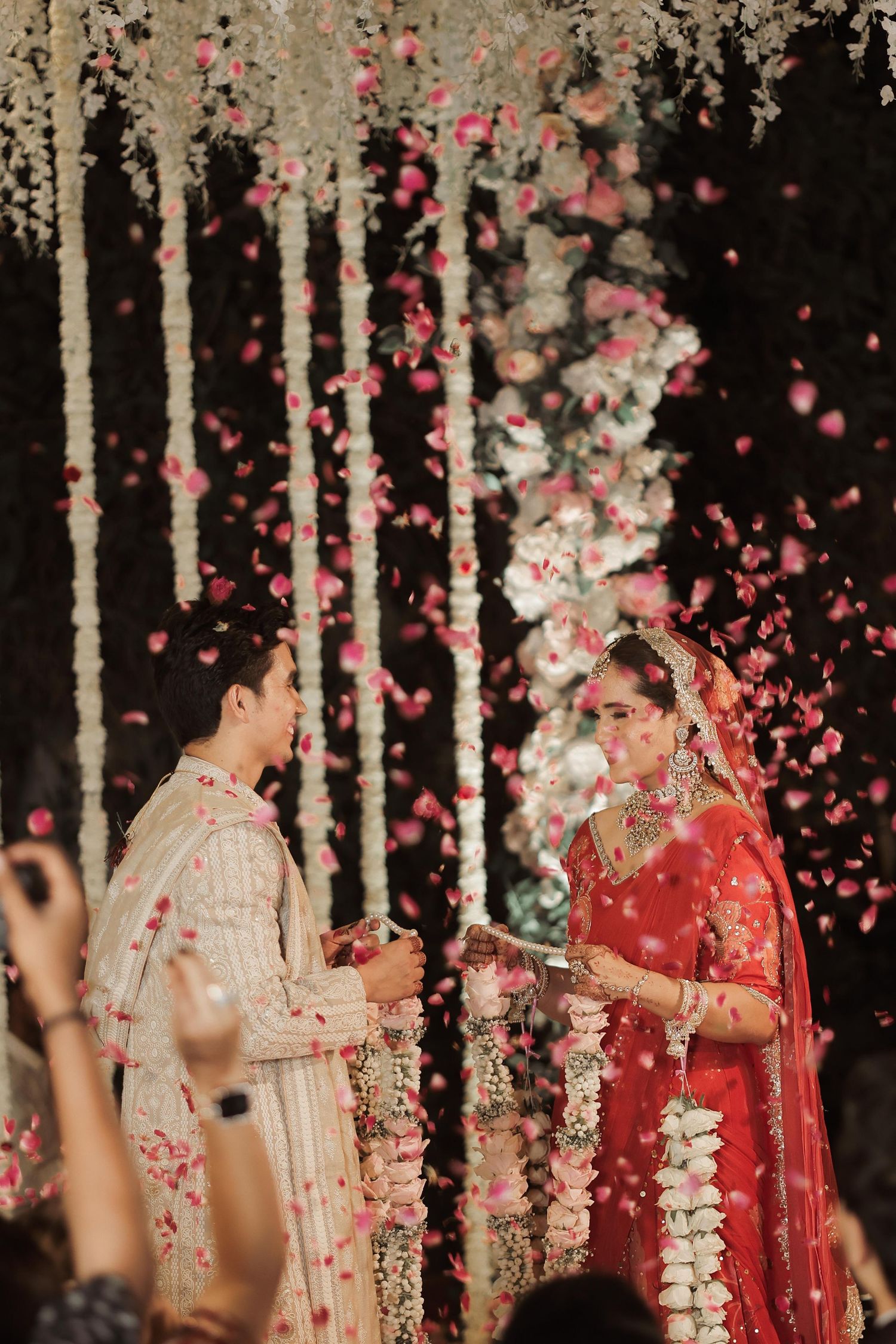 Indian bride in red lehenga and groom exchange garlands at wedding ceremony with pink flower petals falling.