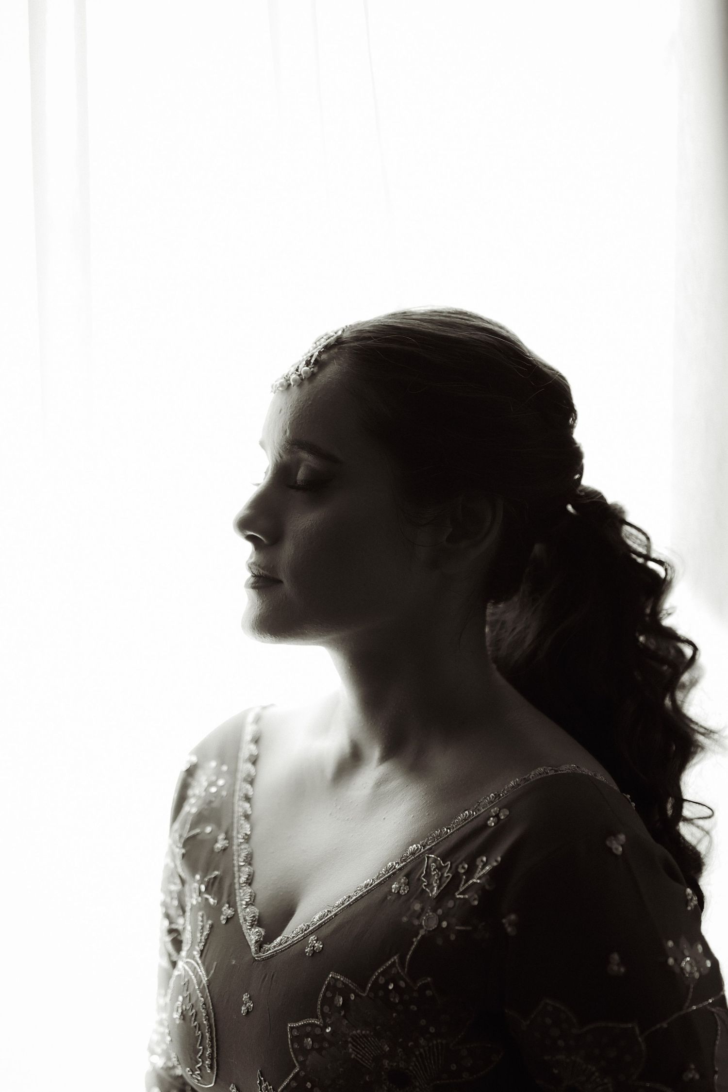 Black and white profile portrait of an elegant bride in embroidered gown with curly ponytail against bright window light.