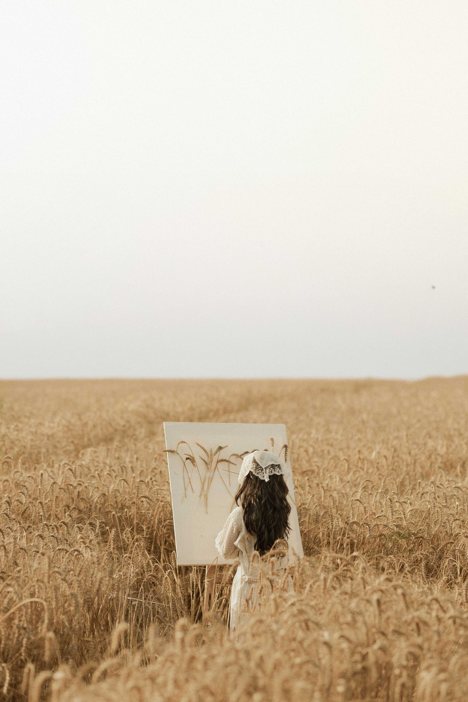 A woman in a white dress holds a canvas painting in a golden wheat field under a pale sky.