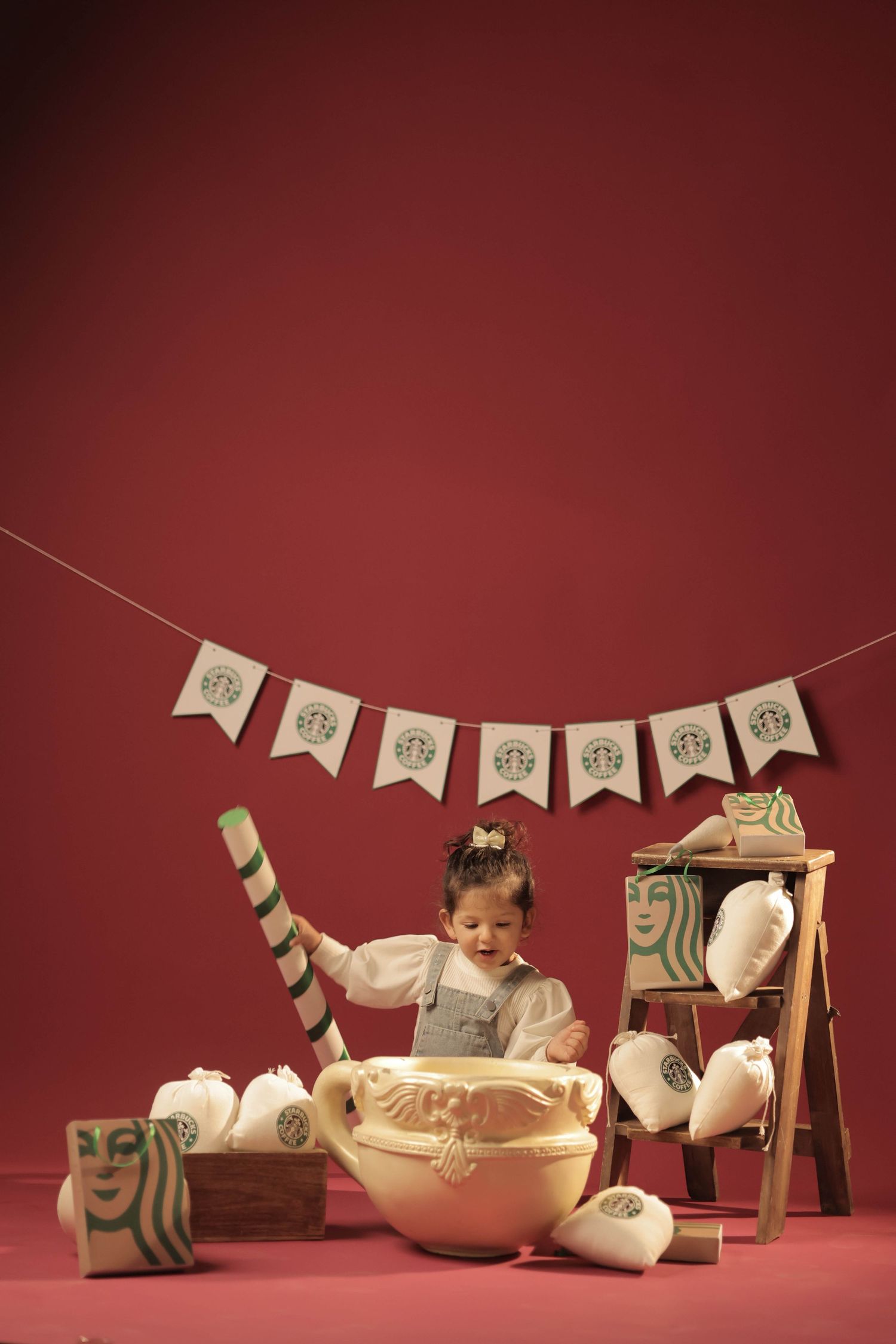 A toddler girl sits in a giant Starbucks-themed bowl surrounded by branded props on a red background.