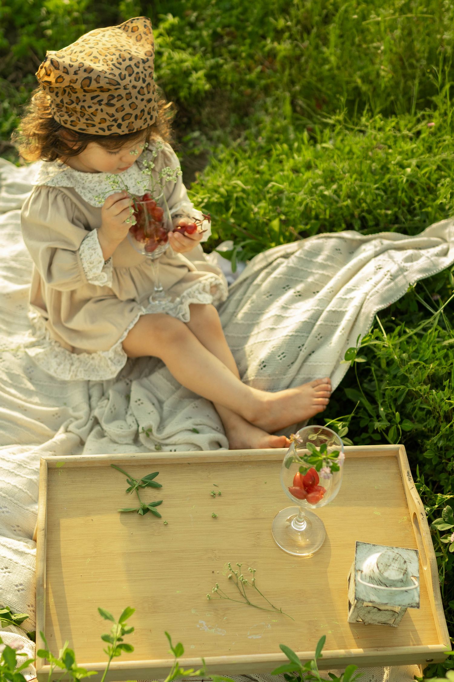 Young girl in vintage dress and leopard hat enjoys a picnic on a blanket with strawberries and a wooden tray.
