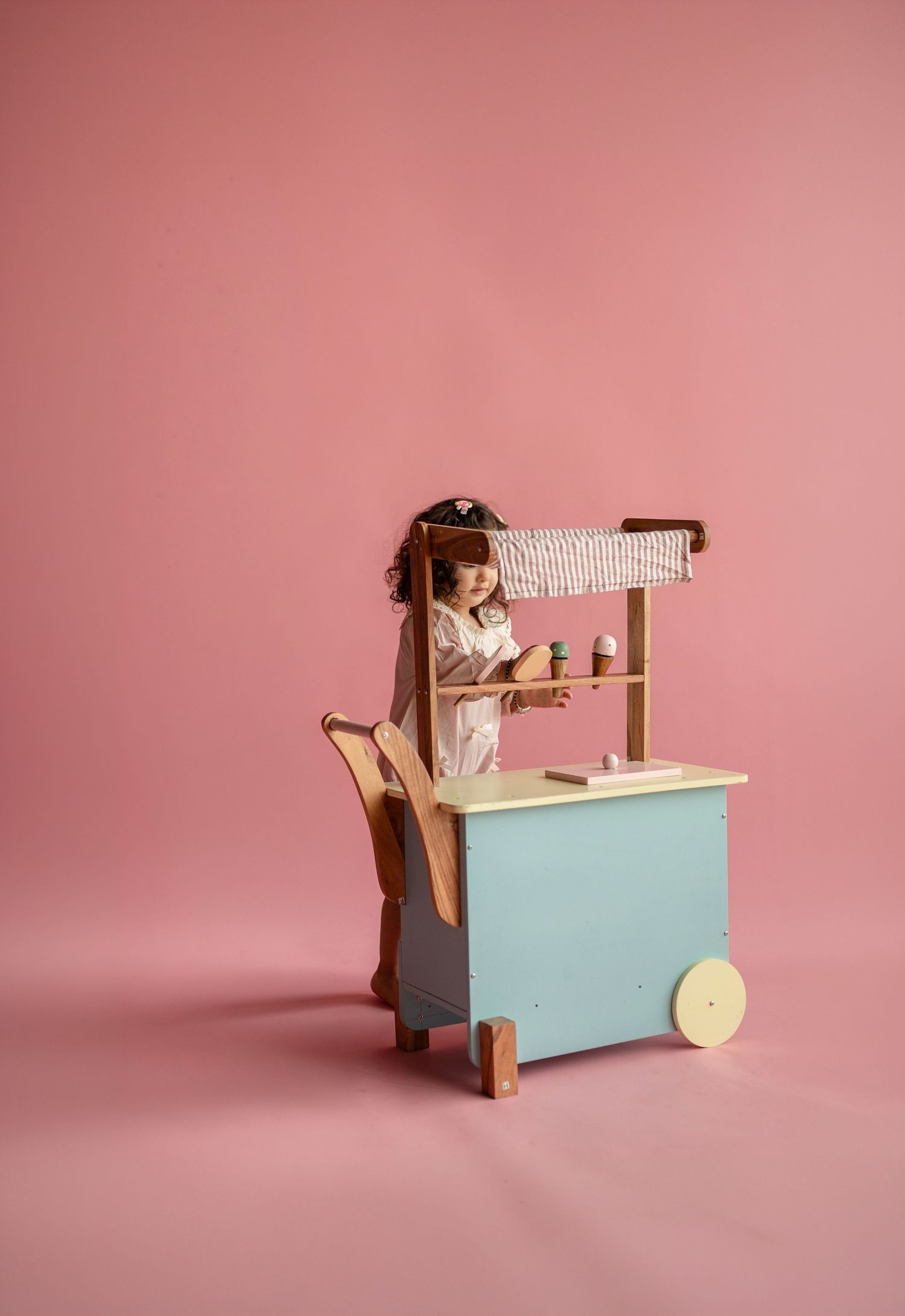 Young girl playing with a wooden toy ice cream cart with blue base and striped canopy on pink background.