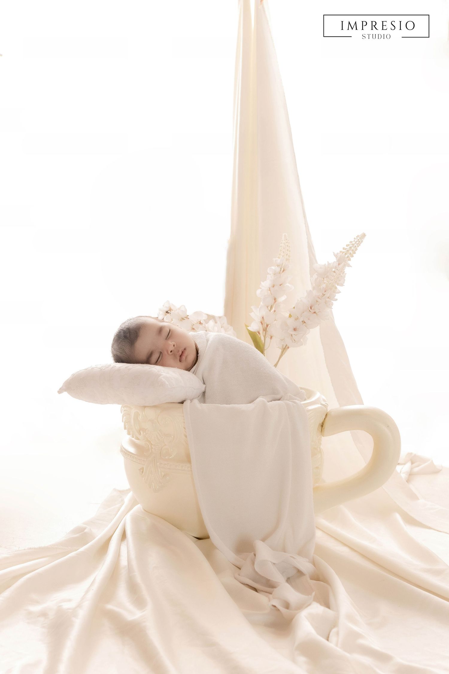Newborn baby sleeping peacefully inside a giant teacup prop with white floral decor.
