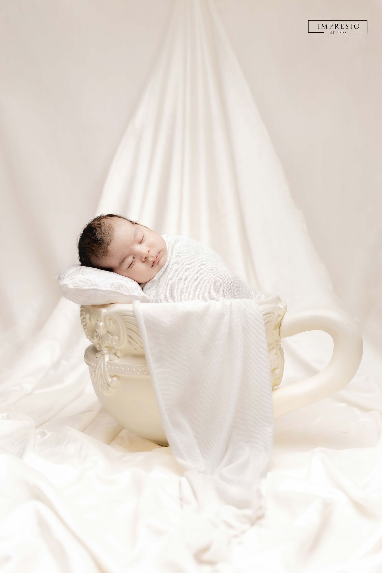 Newborn baby sleeping peacefully wrapped in white swaddle inside an ornate teacup prop on soft white backdrop.