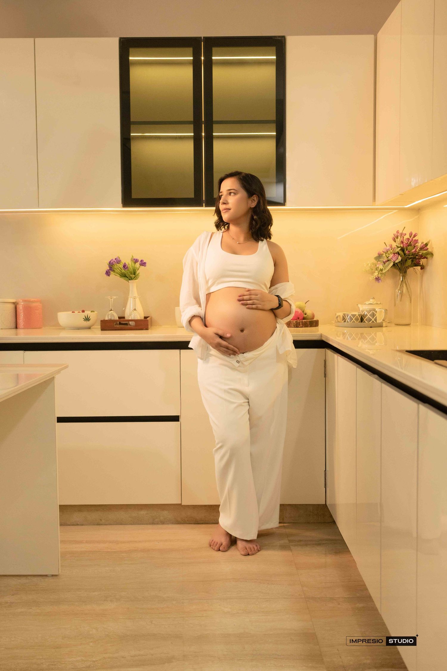 Pregnant woman in white outfit cradling baby bump in modern kitchen during maternity photoshoot.